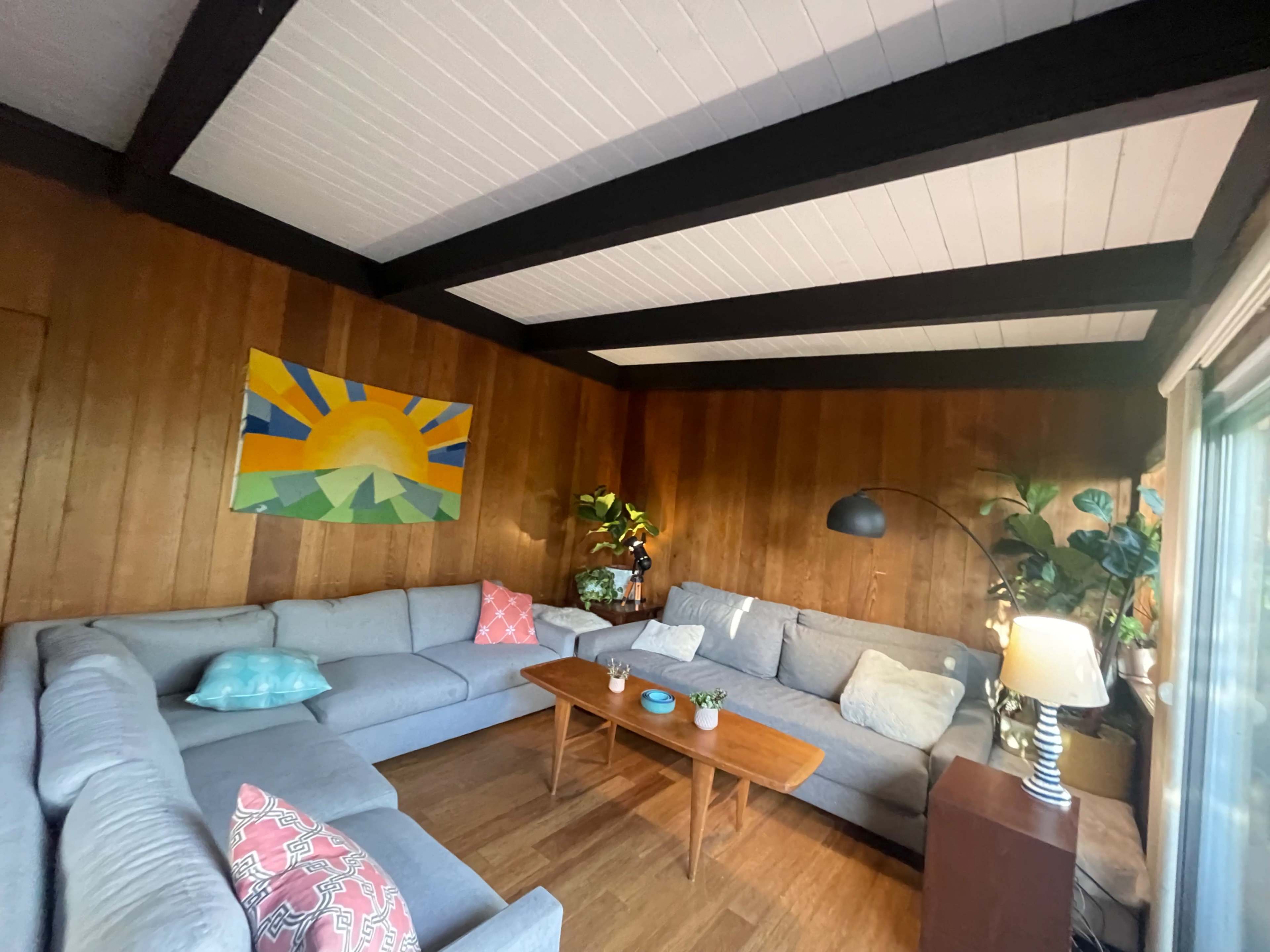 A cozy living room with a gray sectional sofa, a wooden coffee table, and a colorful wall painting, featuring plants and a lamp in the background.