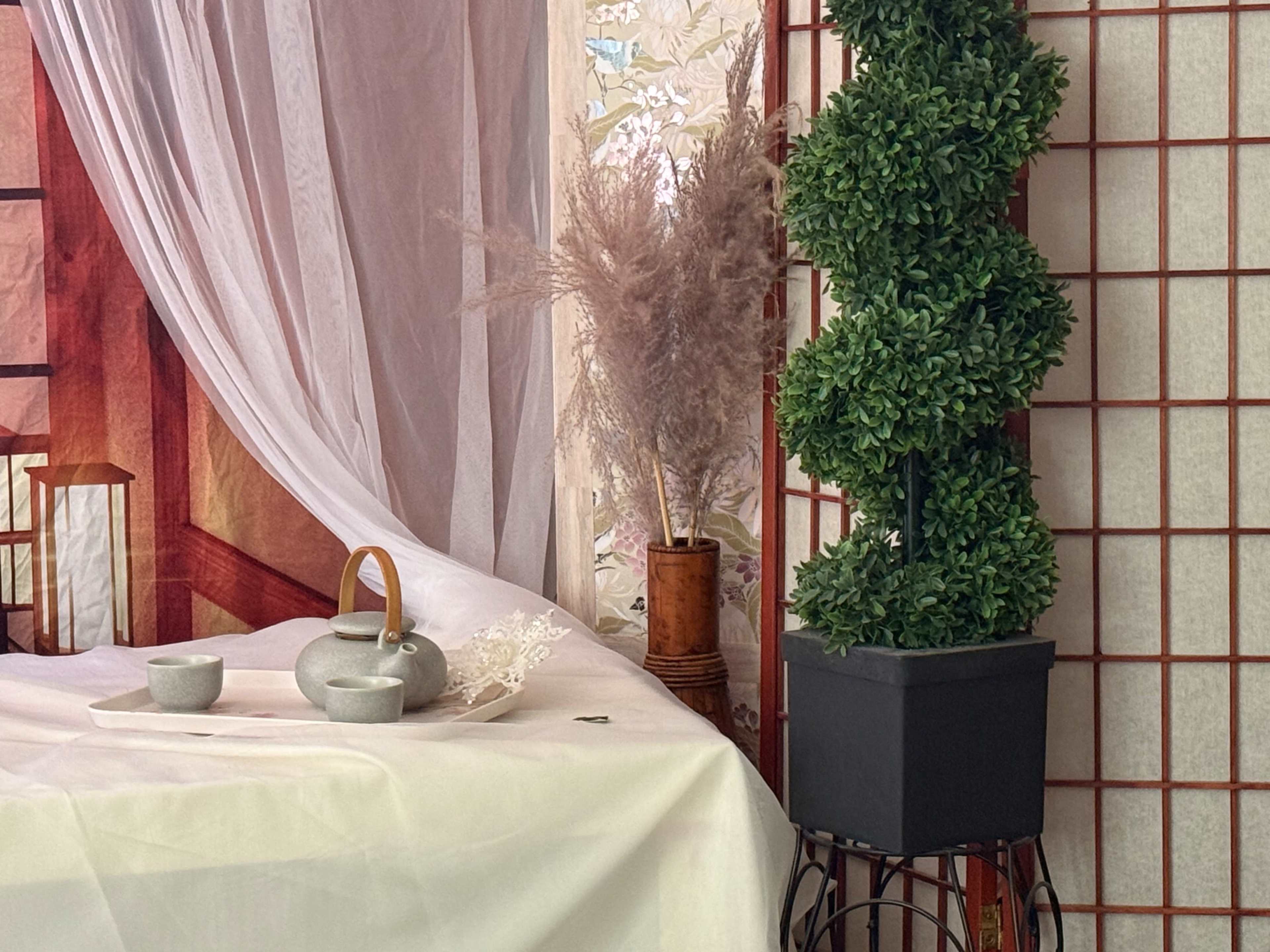 The image shows a decorated interior space featuring a table with a teapot and cups, surrounded by greenery and a decorative screen.