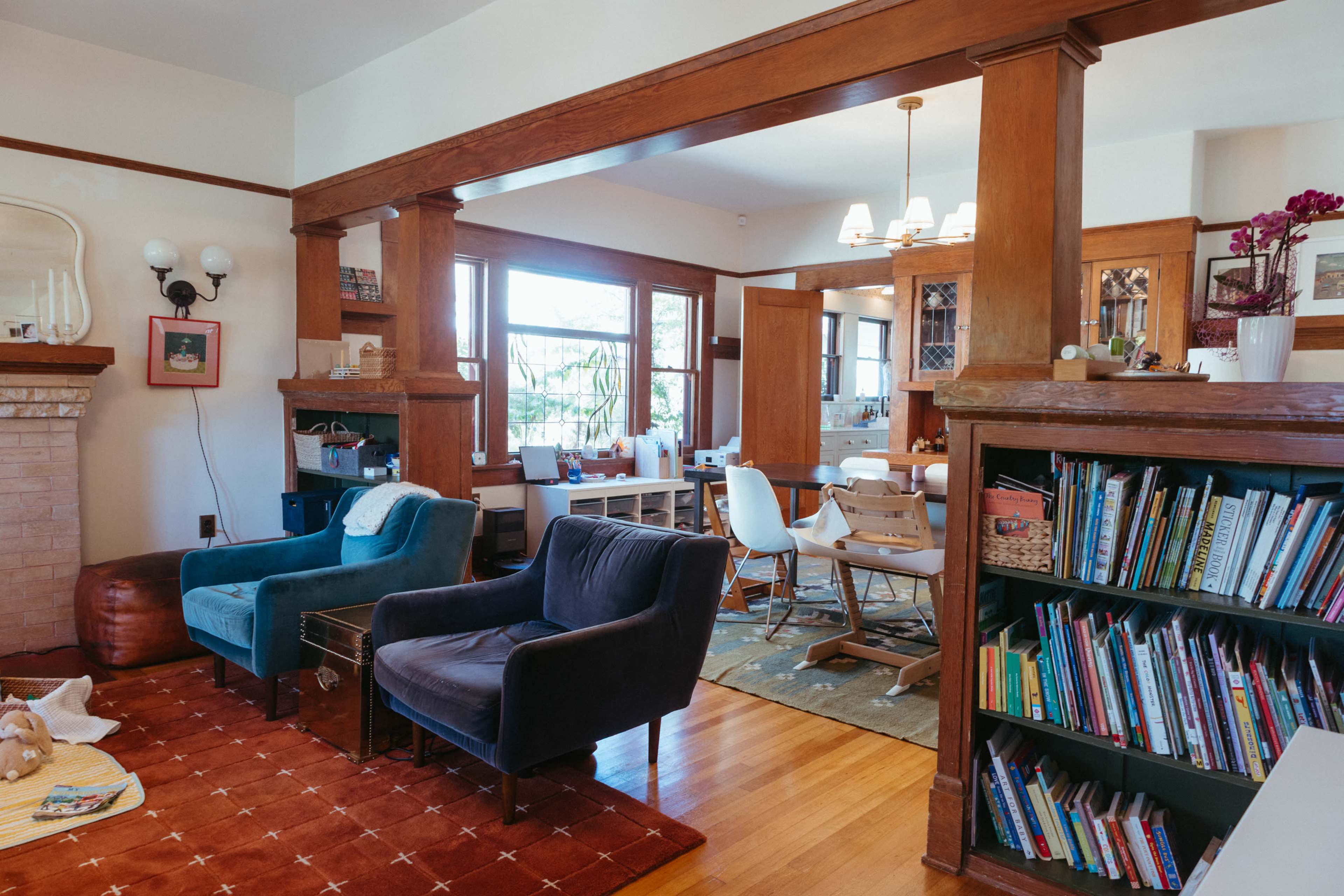 The image shows a cozy living room featuring blue and black armchairs, a bookshelf filled with books, and large windows allowing natural light into the space.