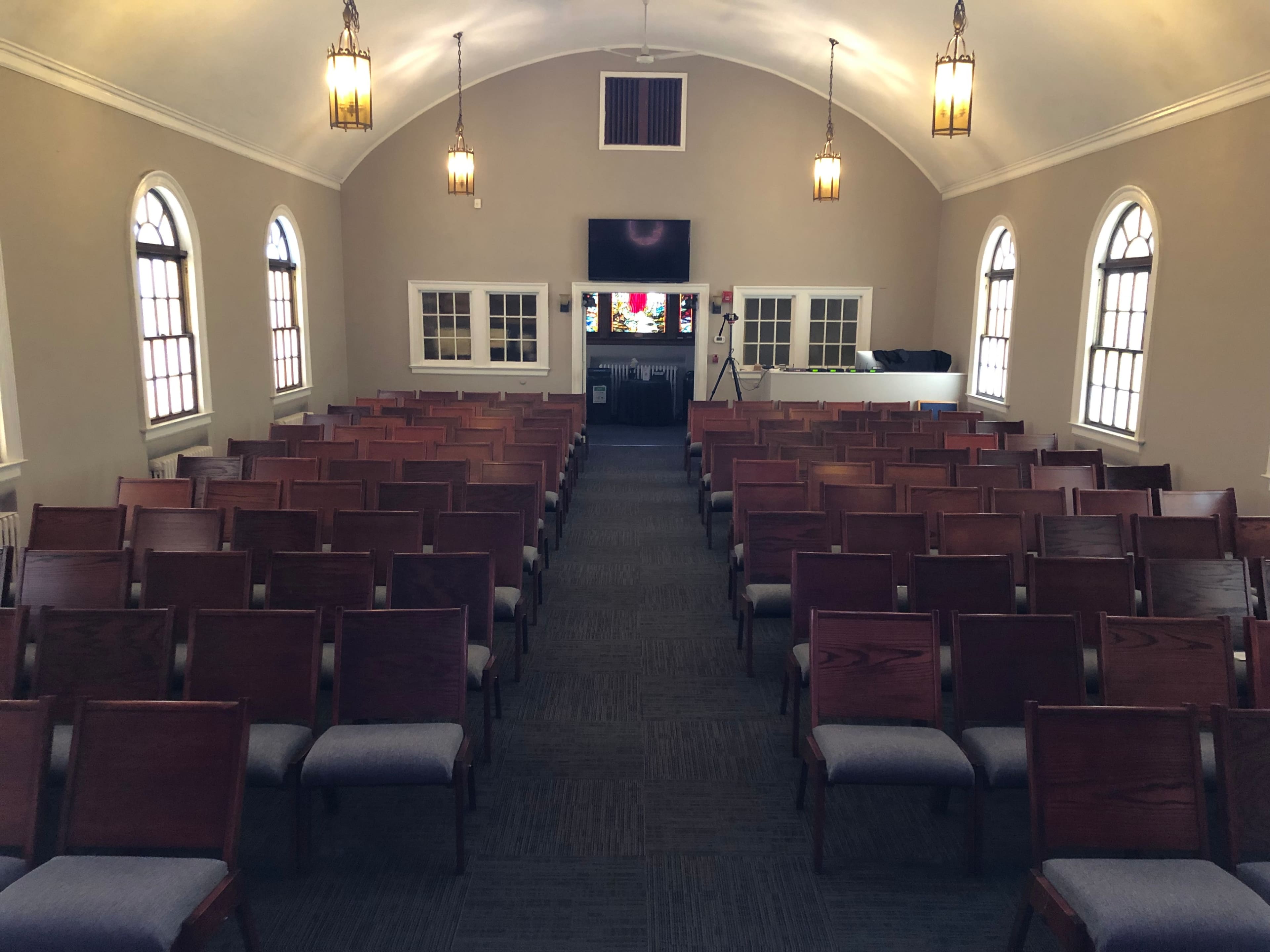 The interior of a church with rows of wooden chairs facing a stage and large windows on either side.