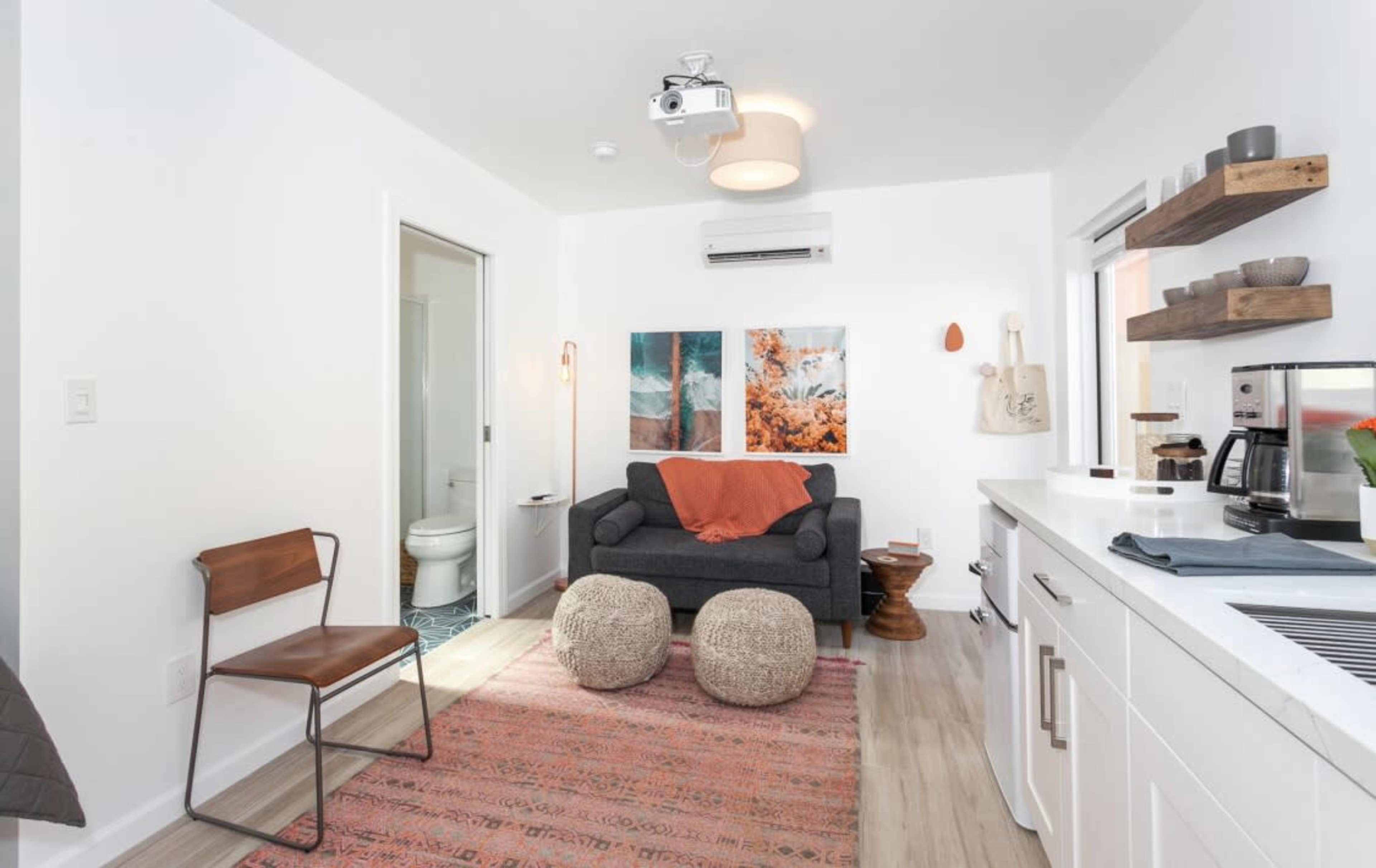 A small living space features a gray couch with an orange throw blanket, a patterned rug, two art pieces on the wall, and a kitchen area with a coffee maker and open shelving.