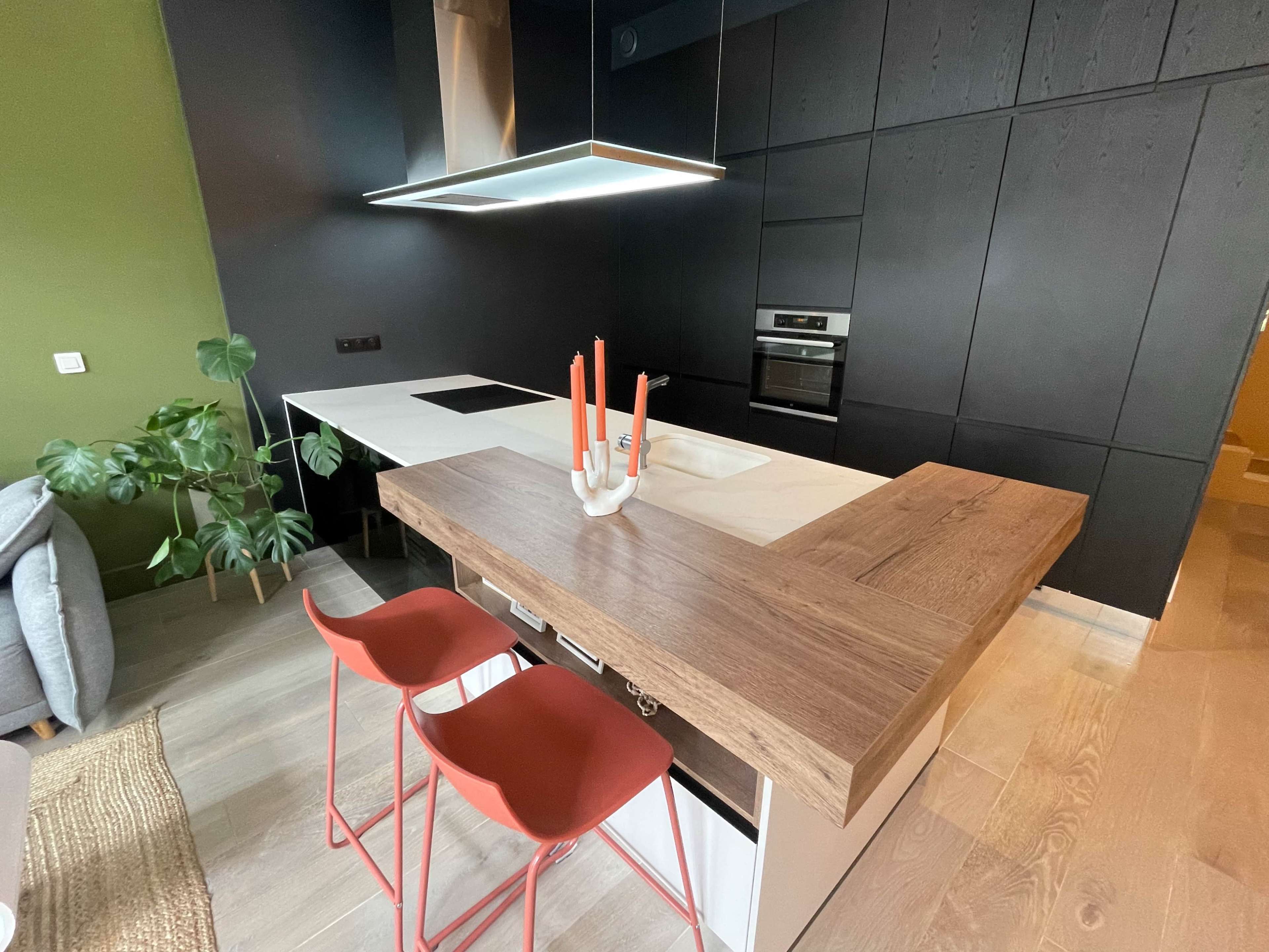 The image shows a modern kitchen with a central island featuring a wooden countertop, red bar stools, and dark cabinetry, complemented by a green accent wall and a potted plant.