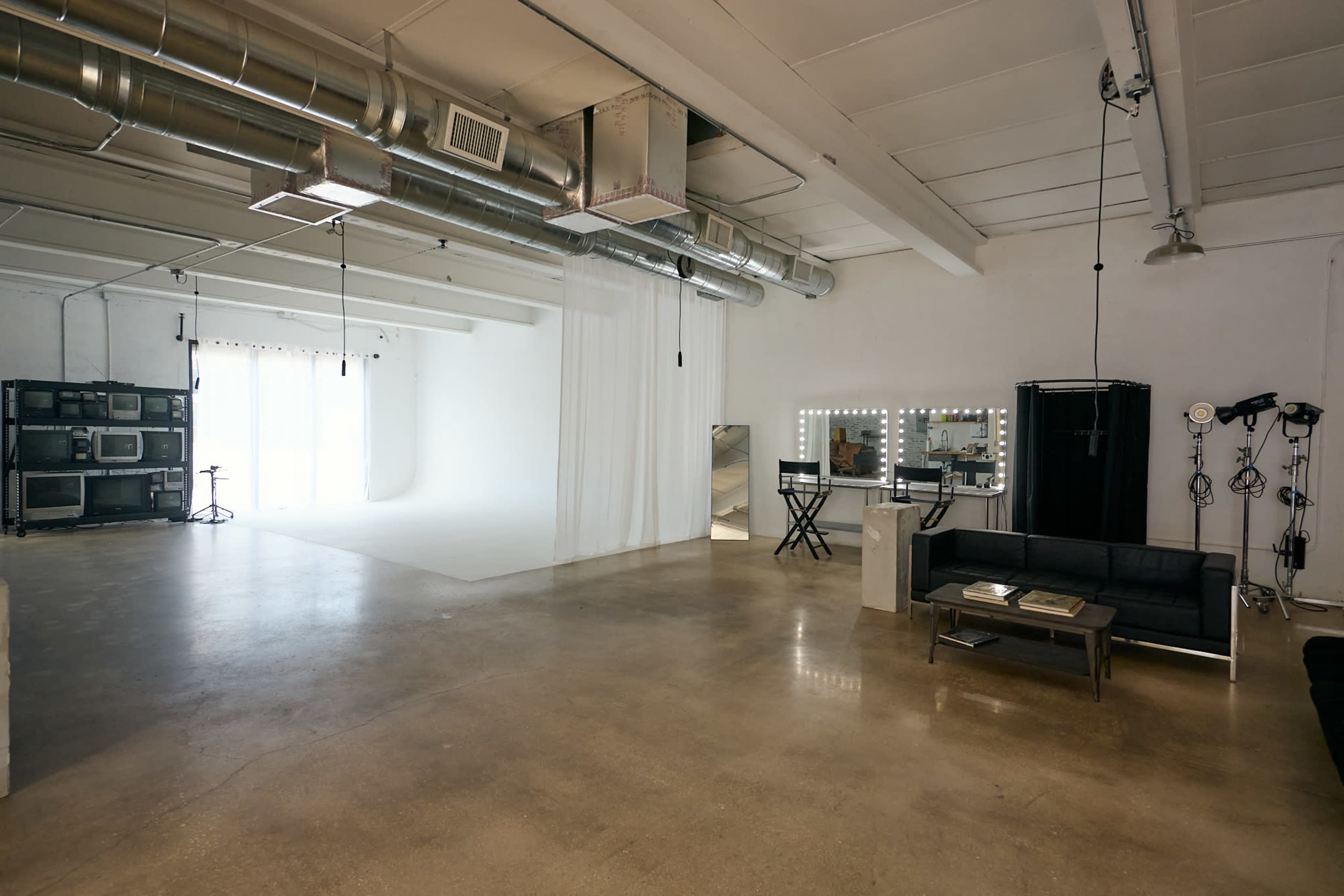 The image shows a spacious studio interior with a white backdrop, a black sofa, and mirrors with bright lights.