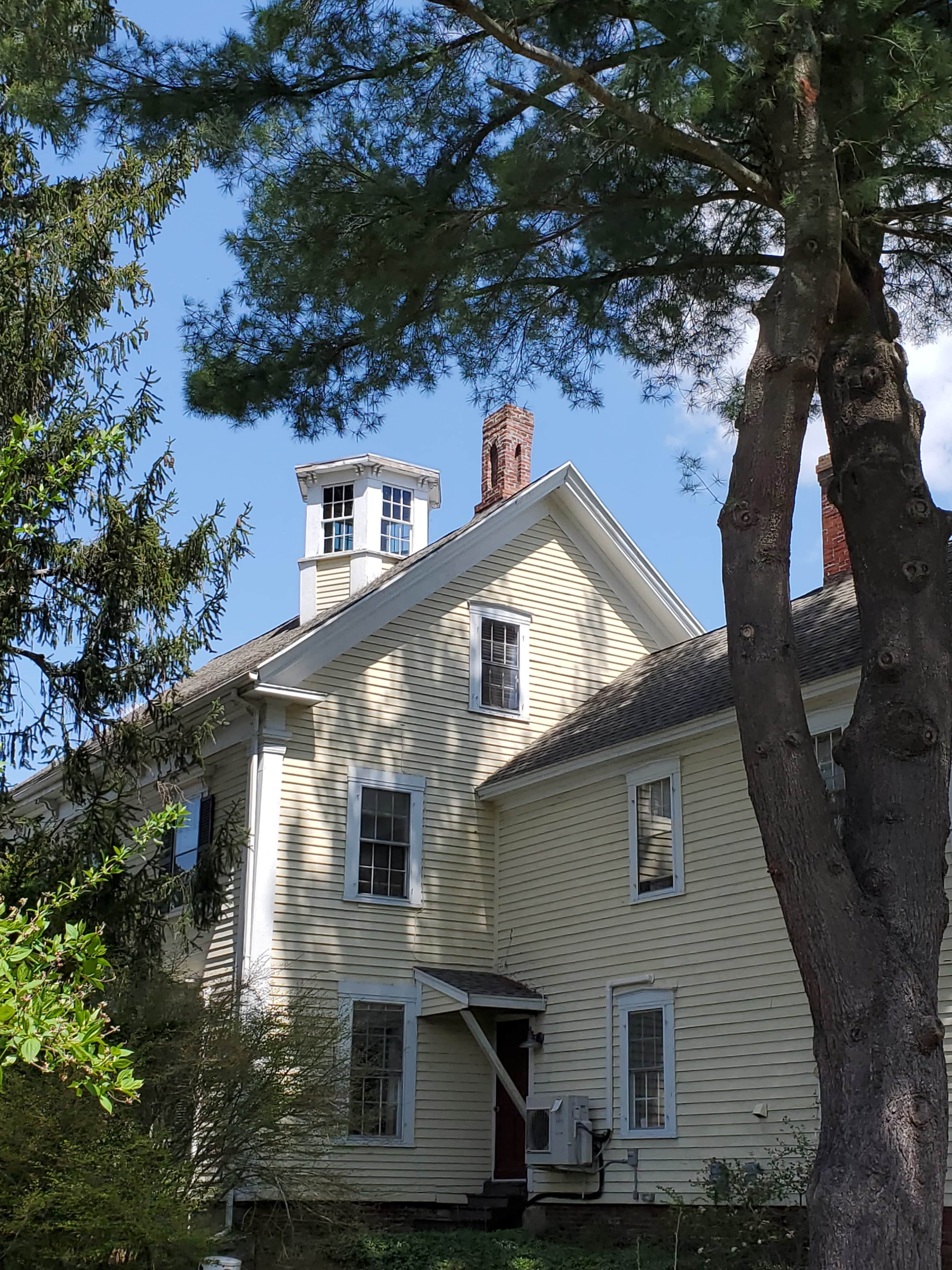 A yellow, two-story house features a brick chimney and a gabled roof, surrounded by trees.
