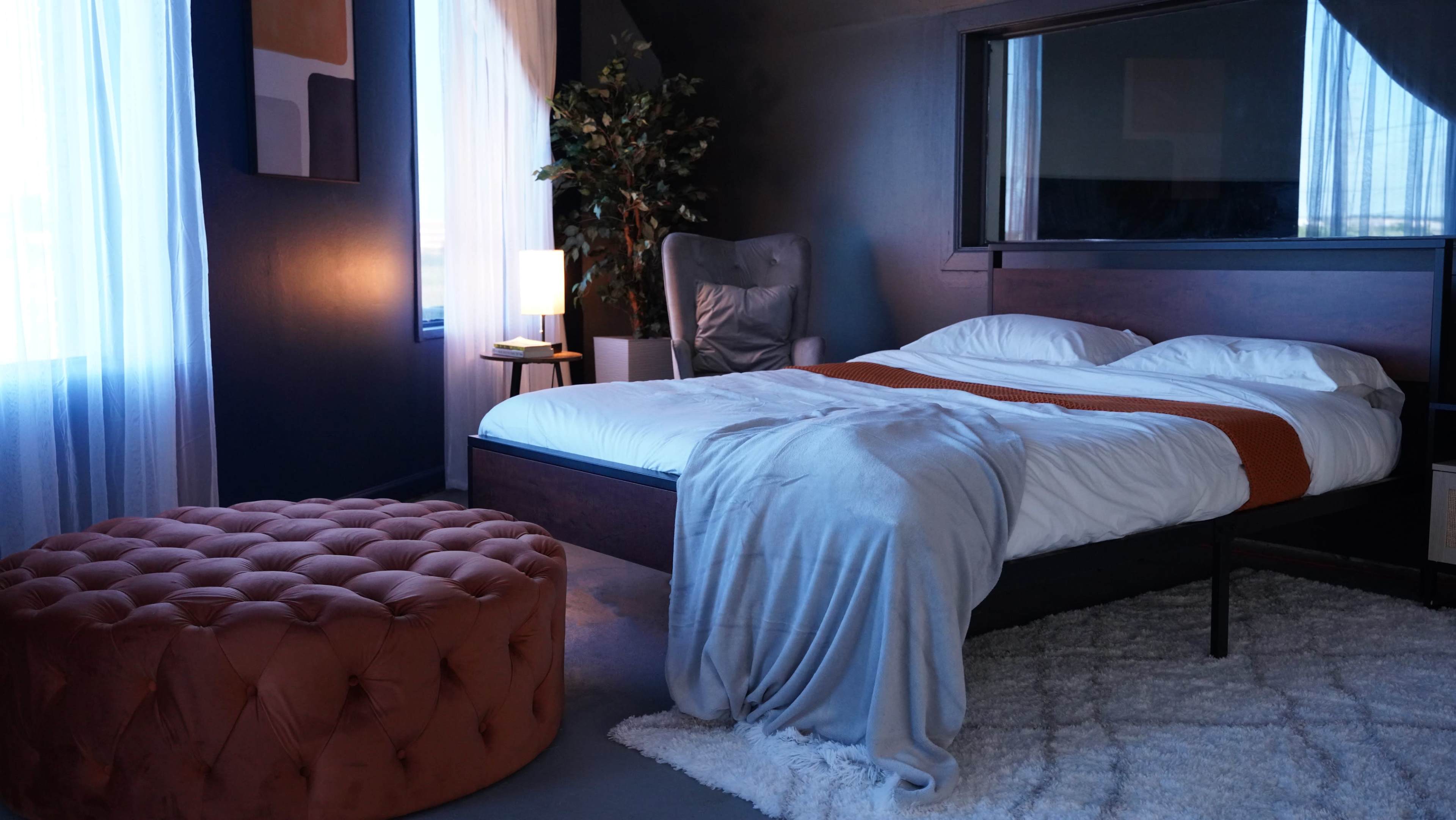 A modern bedroom features a bed with white linens, a tufted ottoman, and large windows allowing natural light to enter.
