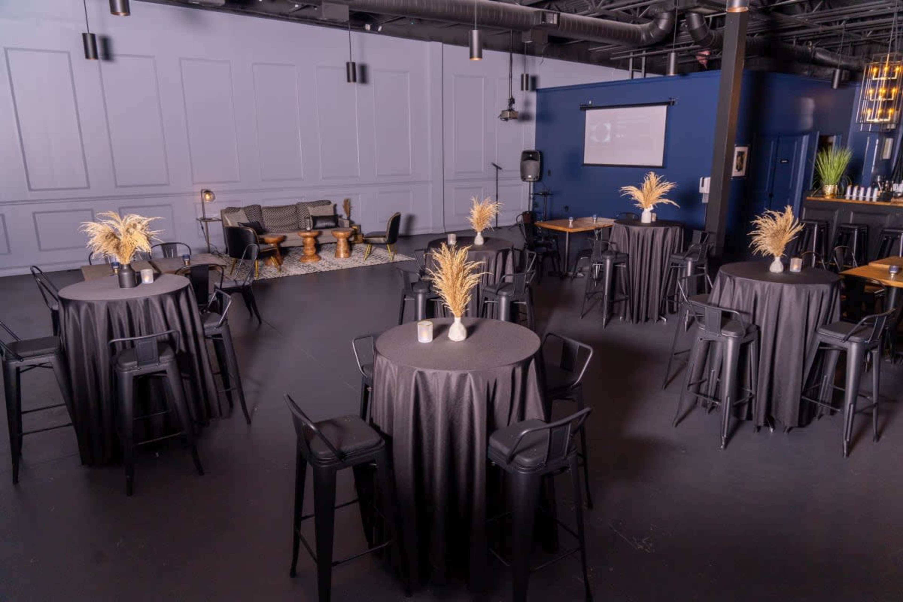 A modern event space with several black tables, decorative plants, and a cozy seating area against a blue wall.
