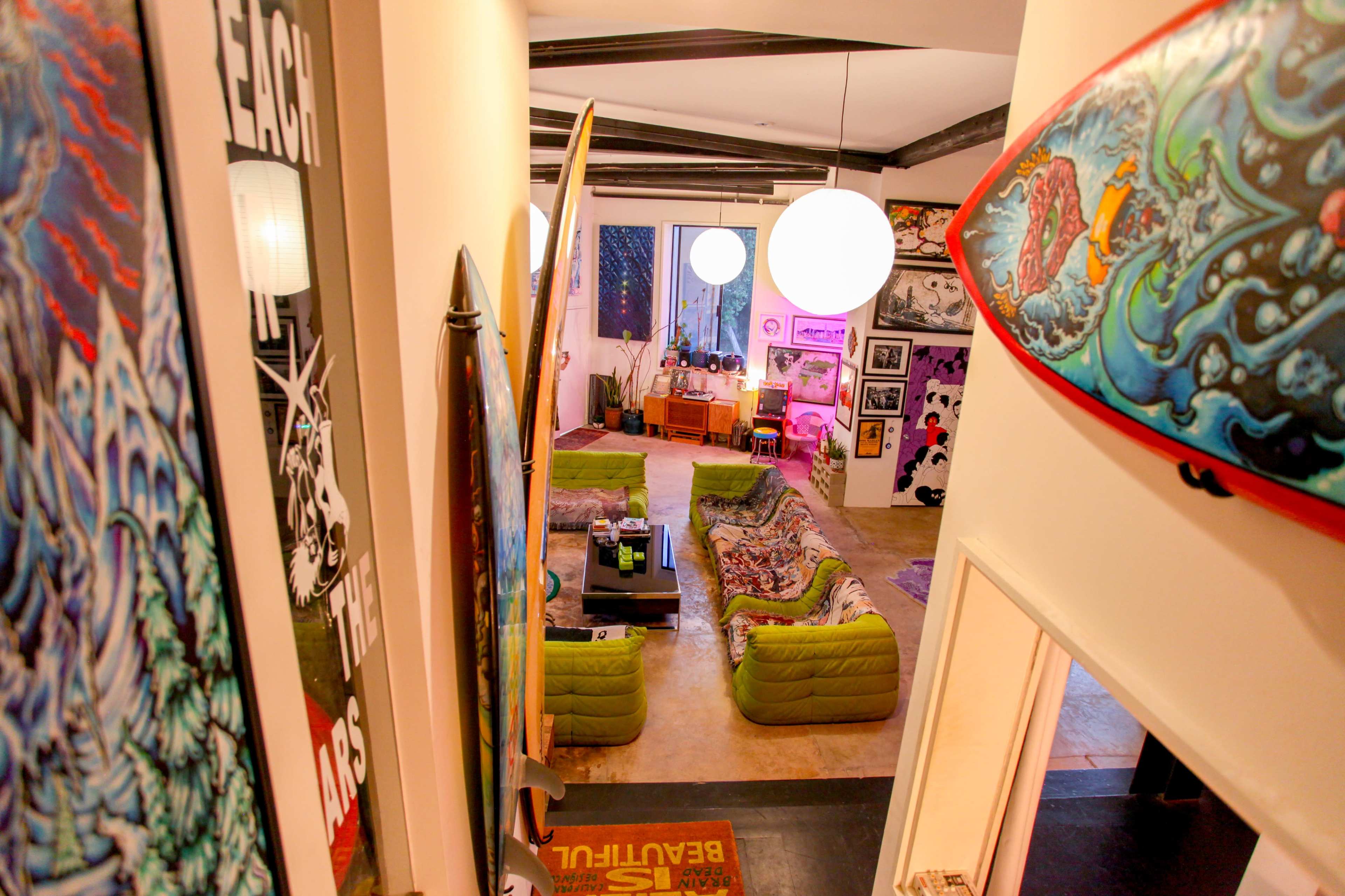 The image shows a colorful living room with green couches, surfboards on the walls, and various artworks displayed.
