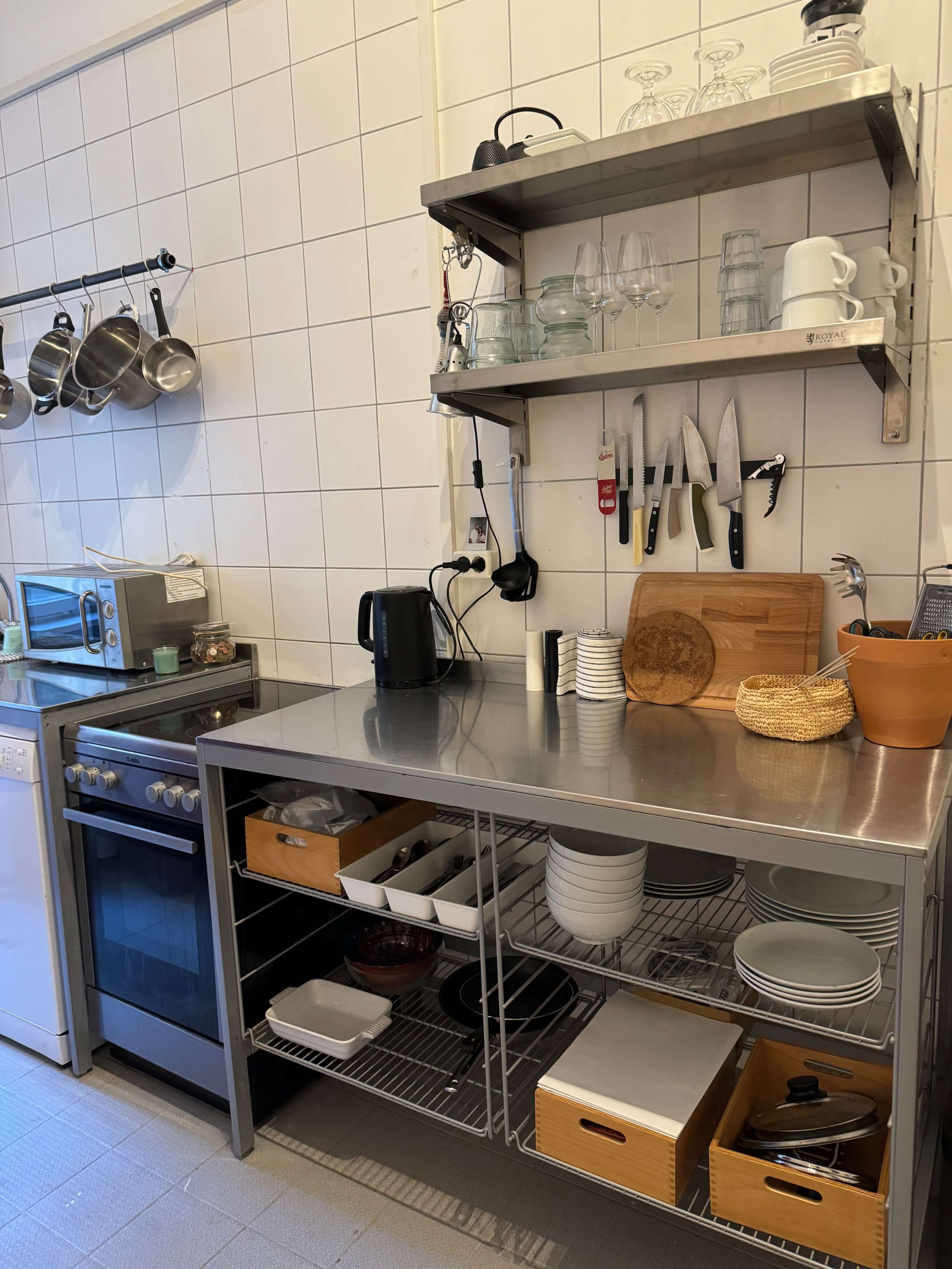 The image shows a stainless steel kitchen workspace with shelves holding glassware and utensils, along with a stove, microwave, and organized dishware below.