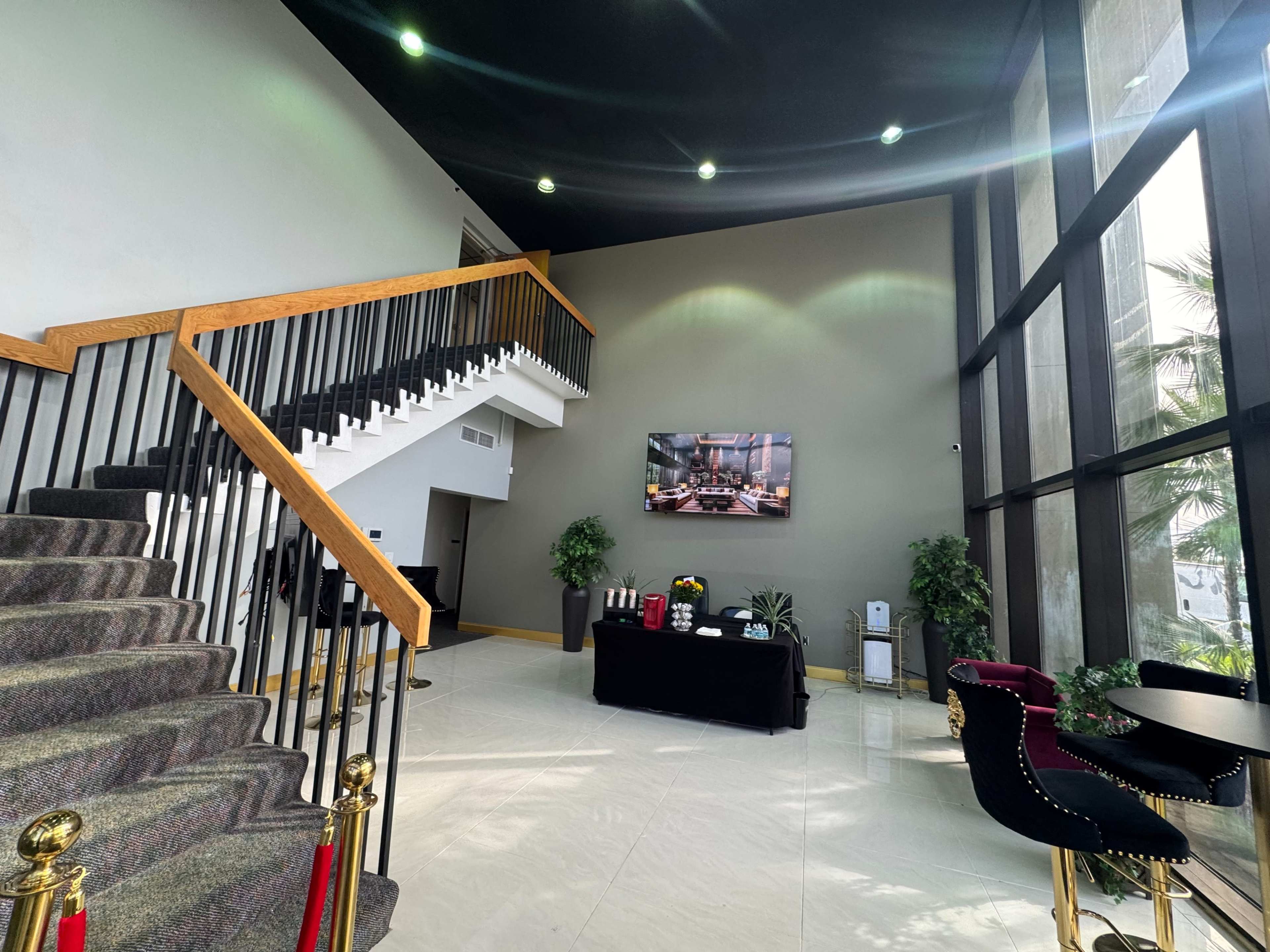 The image shows a spacious lobby with a staircase on the left, a large television mounted on the wall, and a seating area with plants and a table.