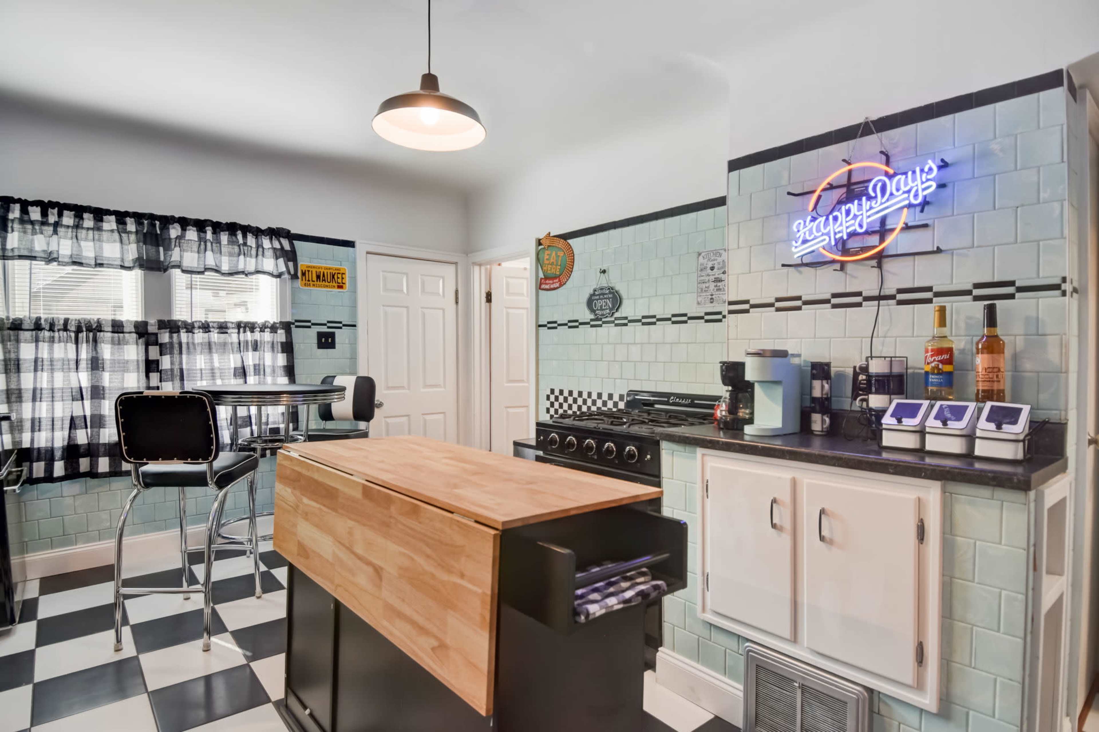 The kitchen features a checkered black-and-white floor, a wooden island, and retro-style decor with blue tiles and a neon sign.