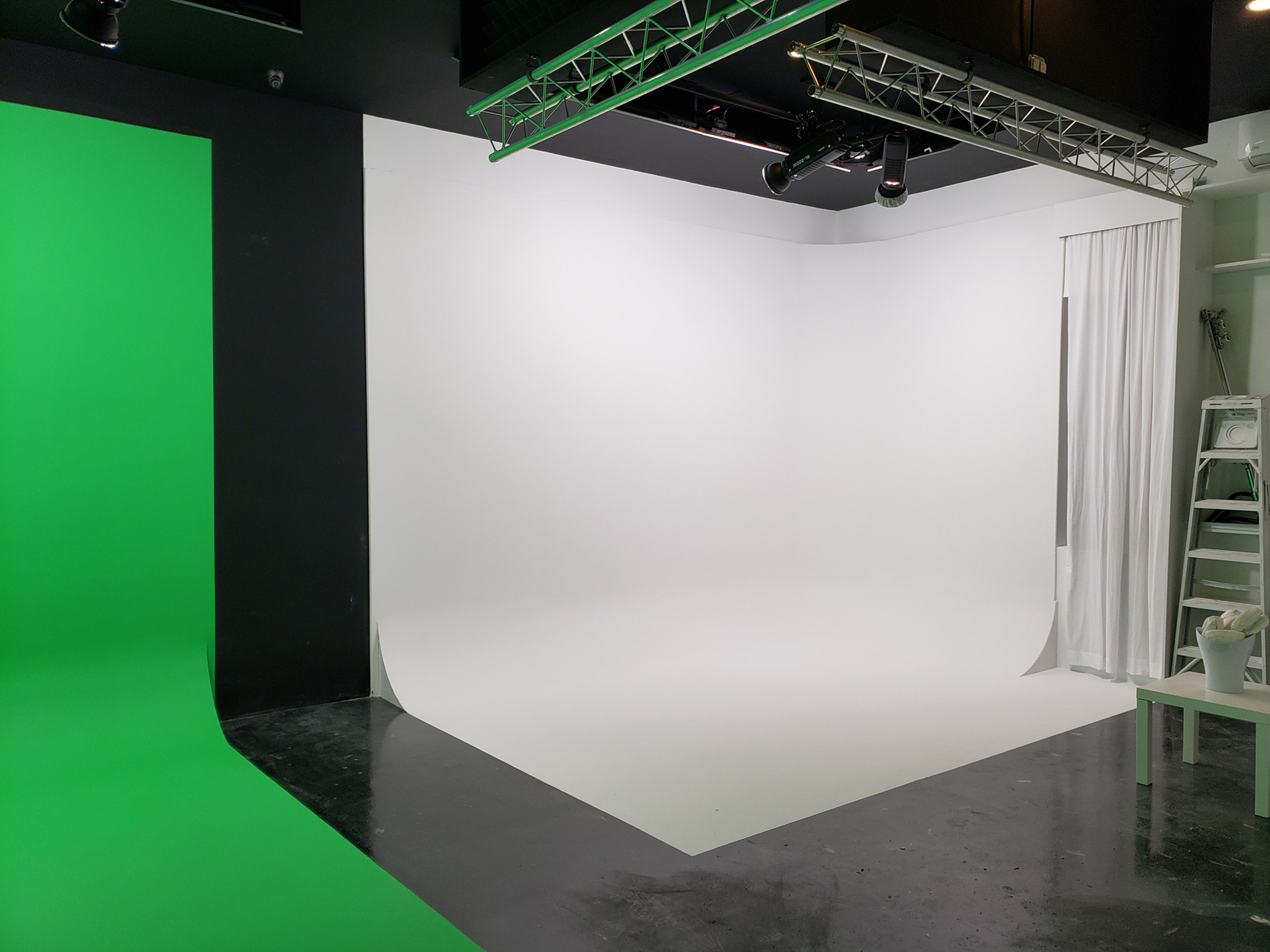 The image shows a photography studio with a green backdrop on one side and a white backdrop on the other, featuring lighting fixtures above.