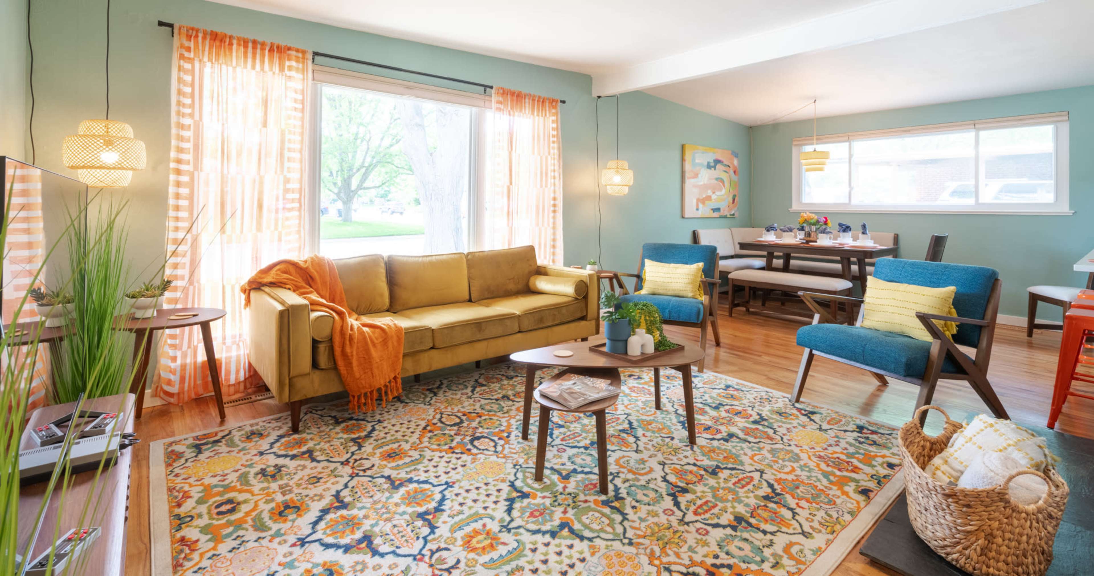 A cozy living room with a yellow sofa, two blue chairs, a patterned rug, and a dining area in the background.