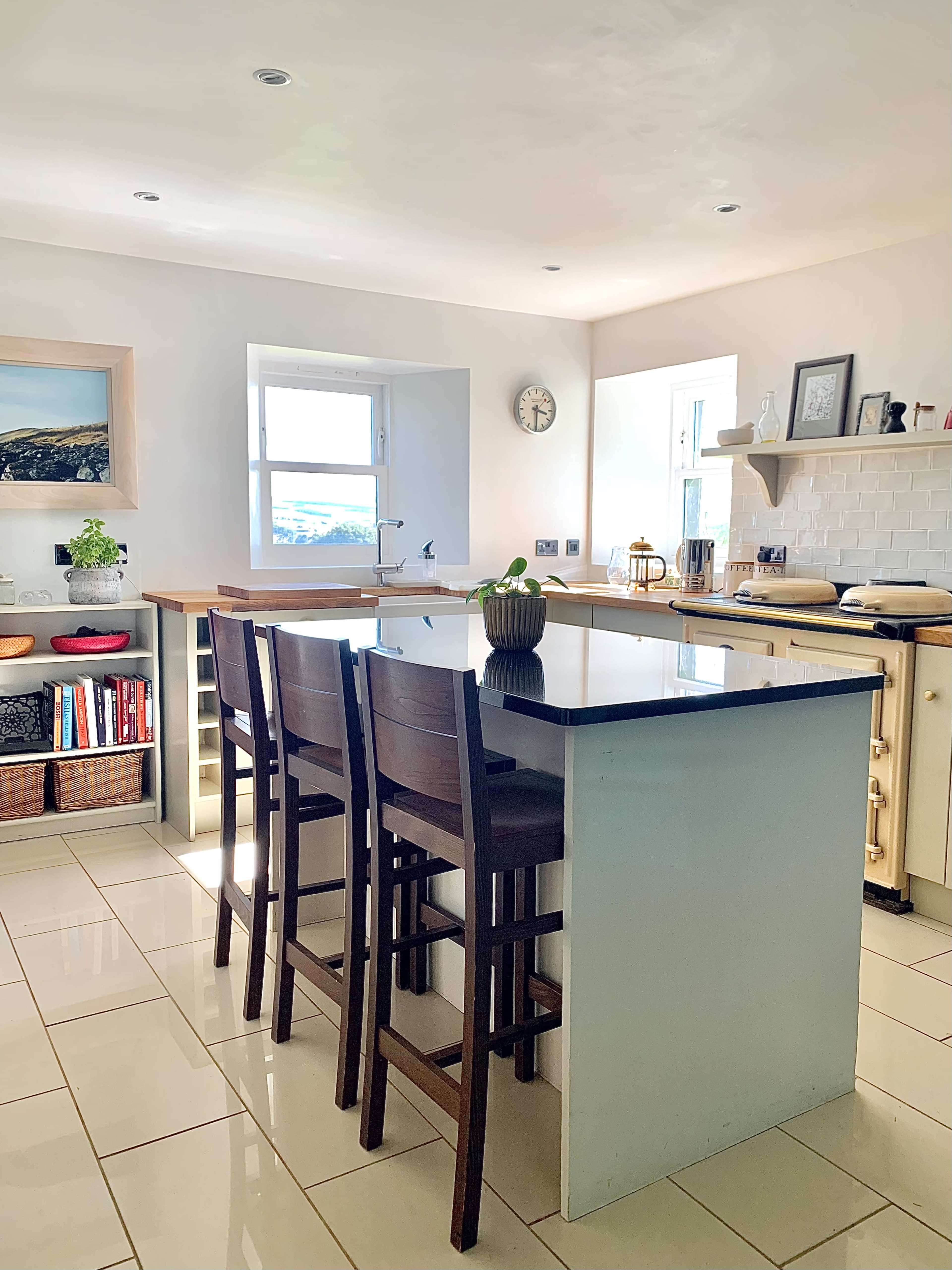 A bright kitchen features a central island with bar stools, a window providing natural light, and organized shelves with dinnerware and decor.