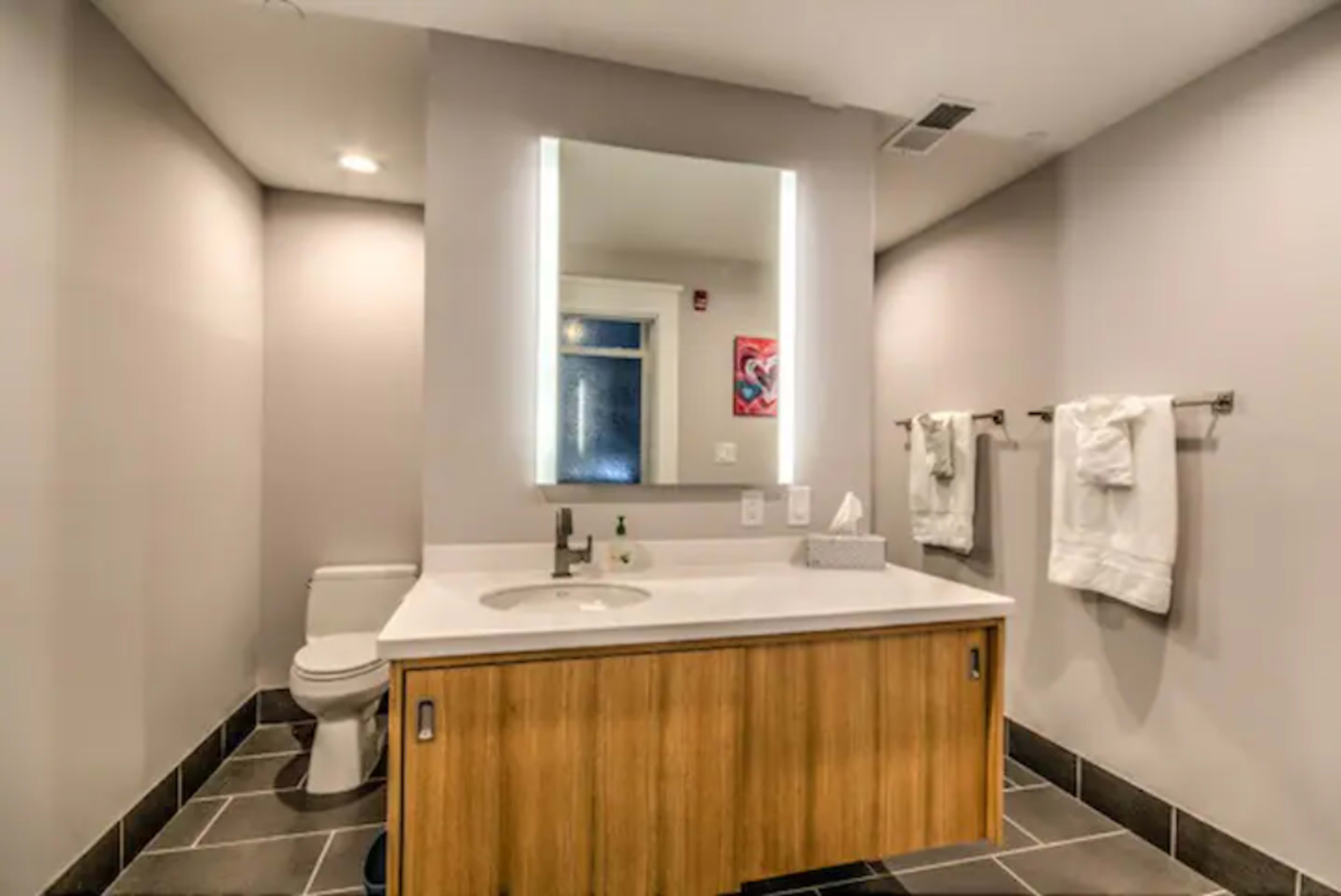 A modern bathroom featuring a wall-mounted mirror above a sink with a wooden vanity, a toilet, and two neatly arranged towels on towel racks.