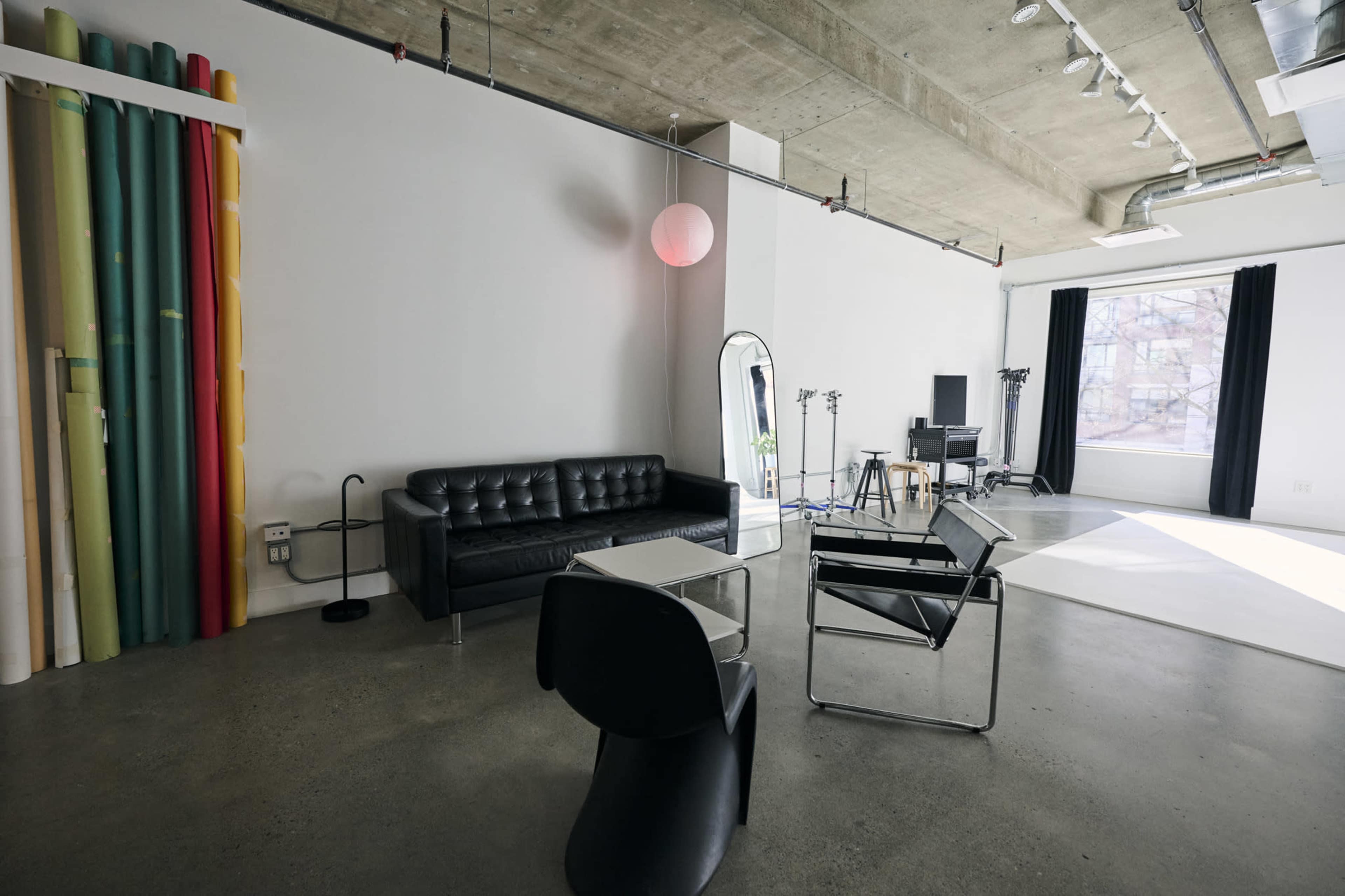The image shows a modern studio space featuring a black leather sofa, a chair, a mirror, and a desk with equipment, along with colorful backdrop rolls on the left and large windows with black curtains on the right.