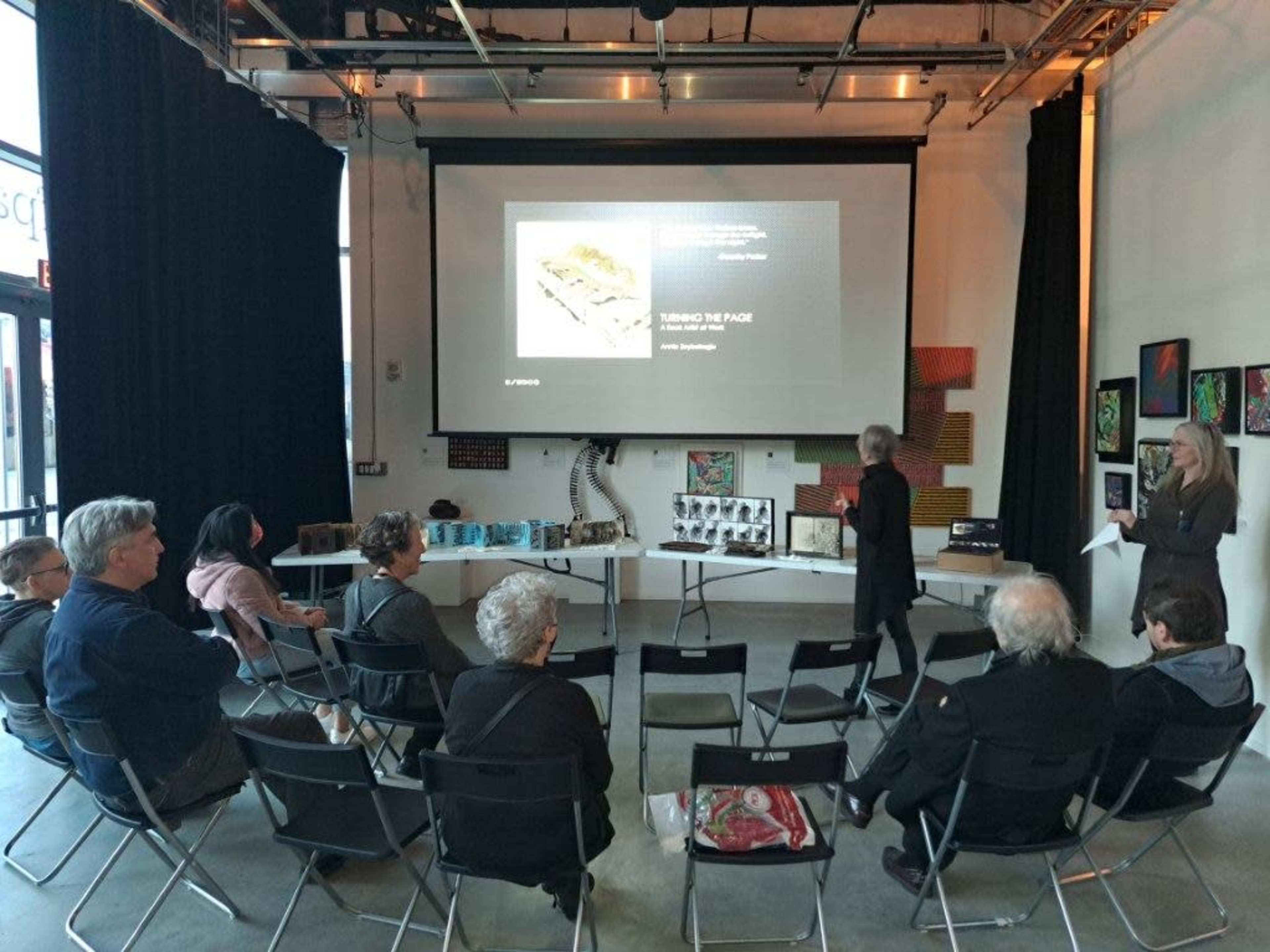 A group of attendees sits in folding chairs facing a screen and a speaker at an event in a well-lit gallery space.