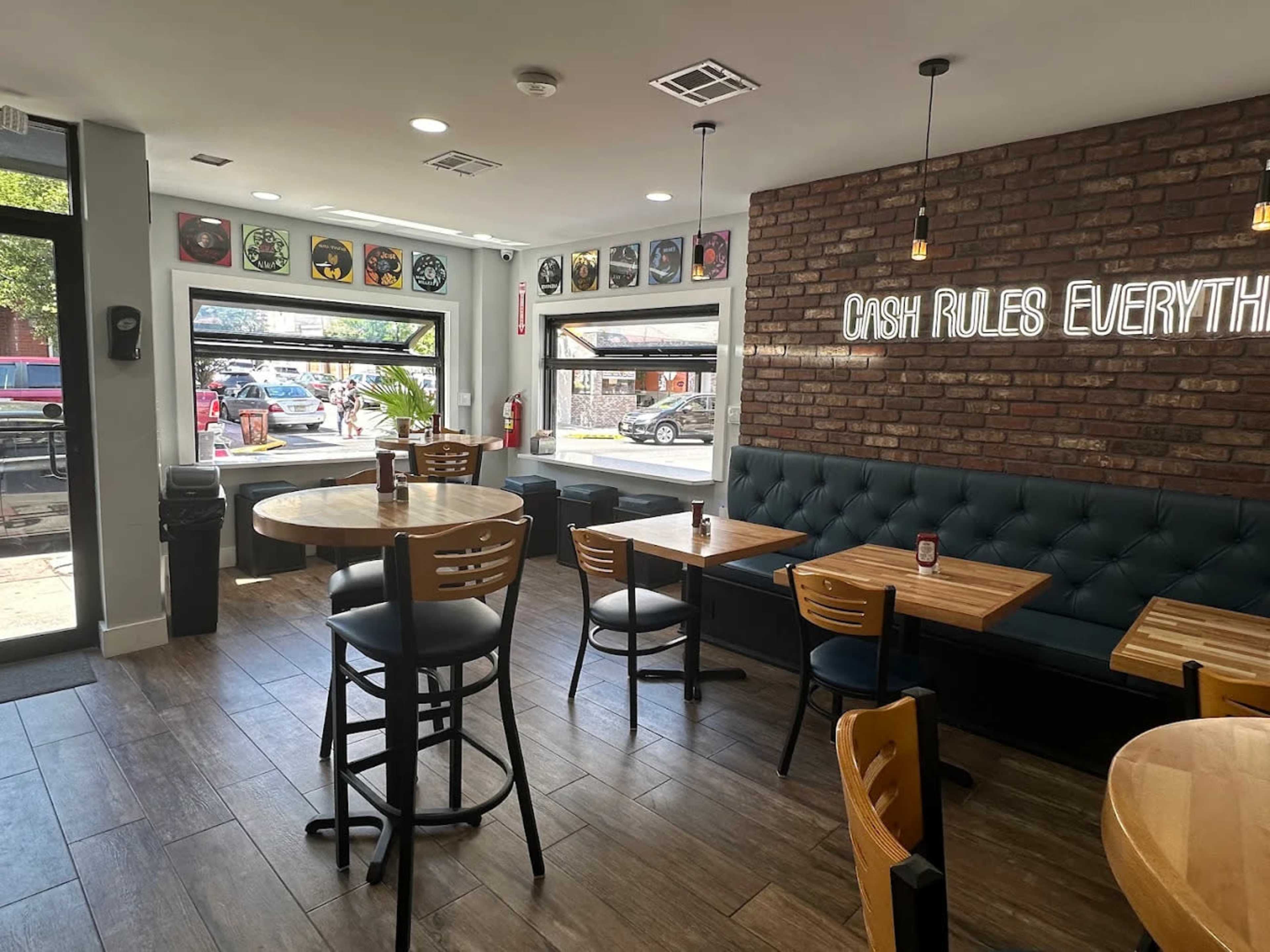The interior of a restaurant features wooden tables and chairs, a brick wall with decorative art, and large windows overlooking a street.