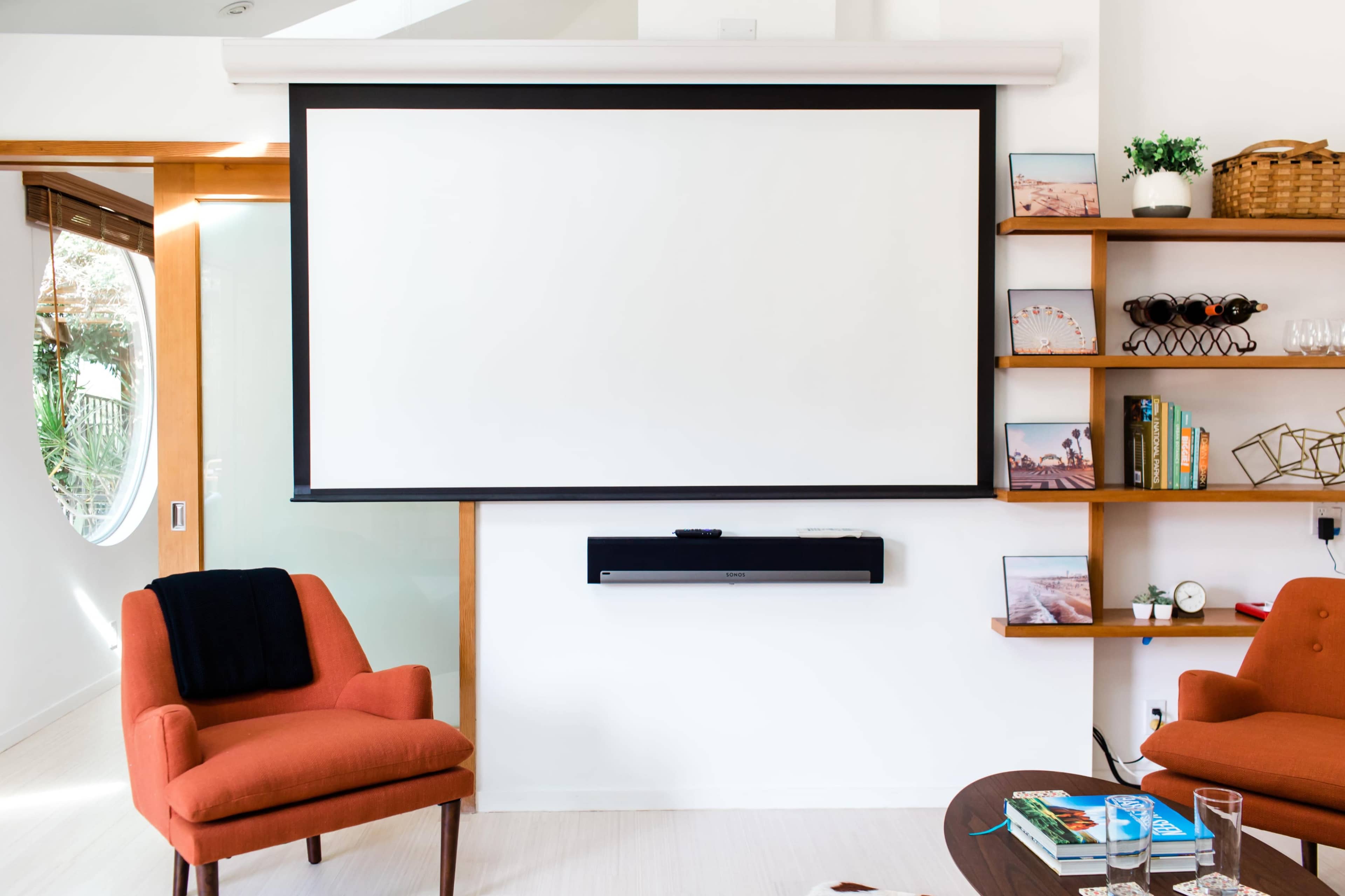 The image shows a cozy living room with two orange chairs facing a wall-mounted projector screen and a soundbar, alongside wooden shelves filled with books and decor.