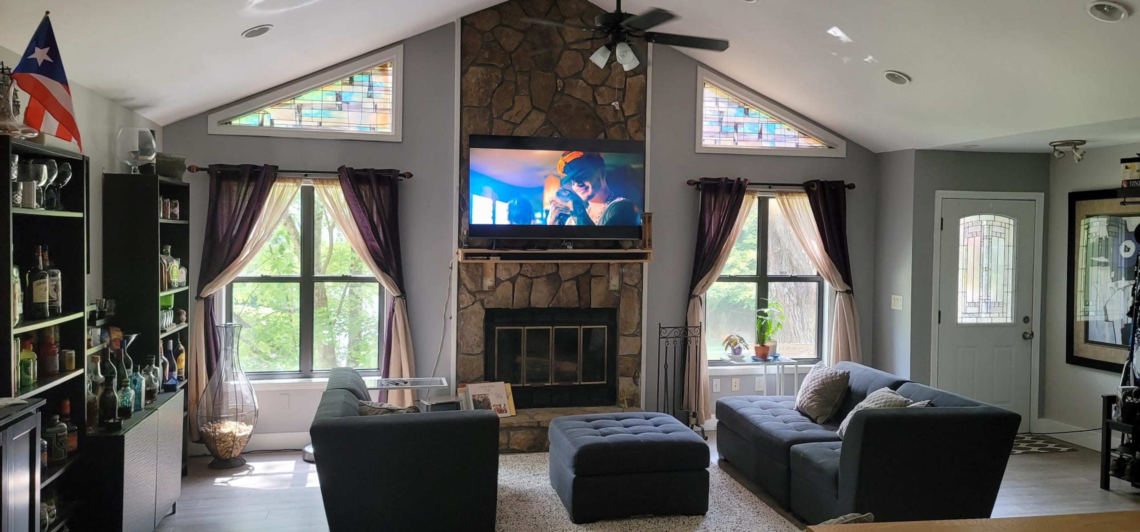 A spacious living room features a large stone fireplace, a mounted television, two dark sofas, and large windows adorned with curtains.