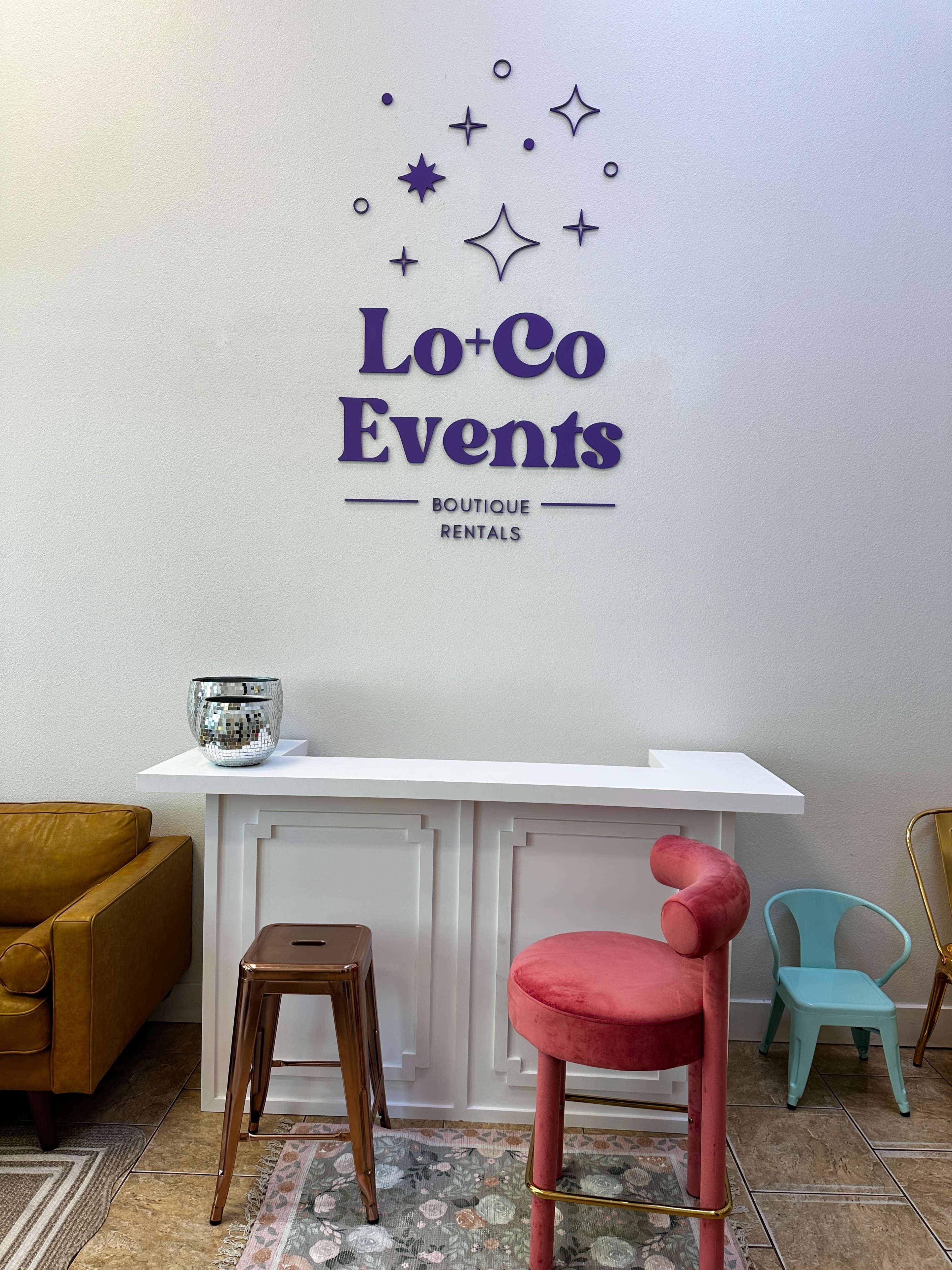 The image shows a reception area of a boutique event rental space, featuring a white counter, colorful seating options, and decorative wall elements.