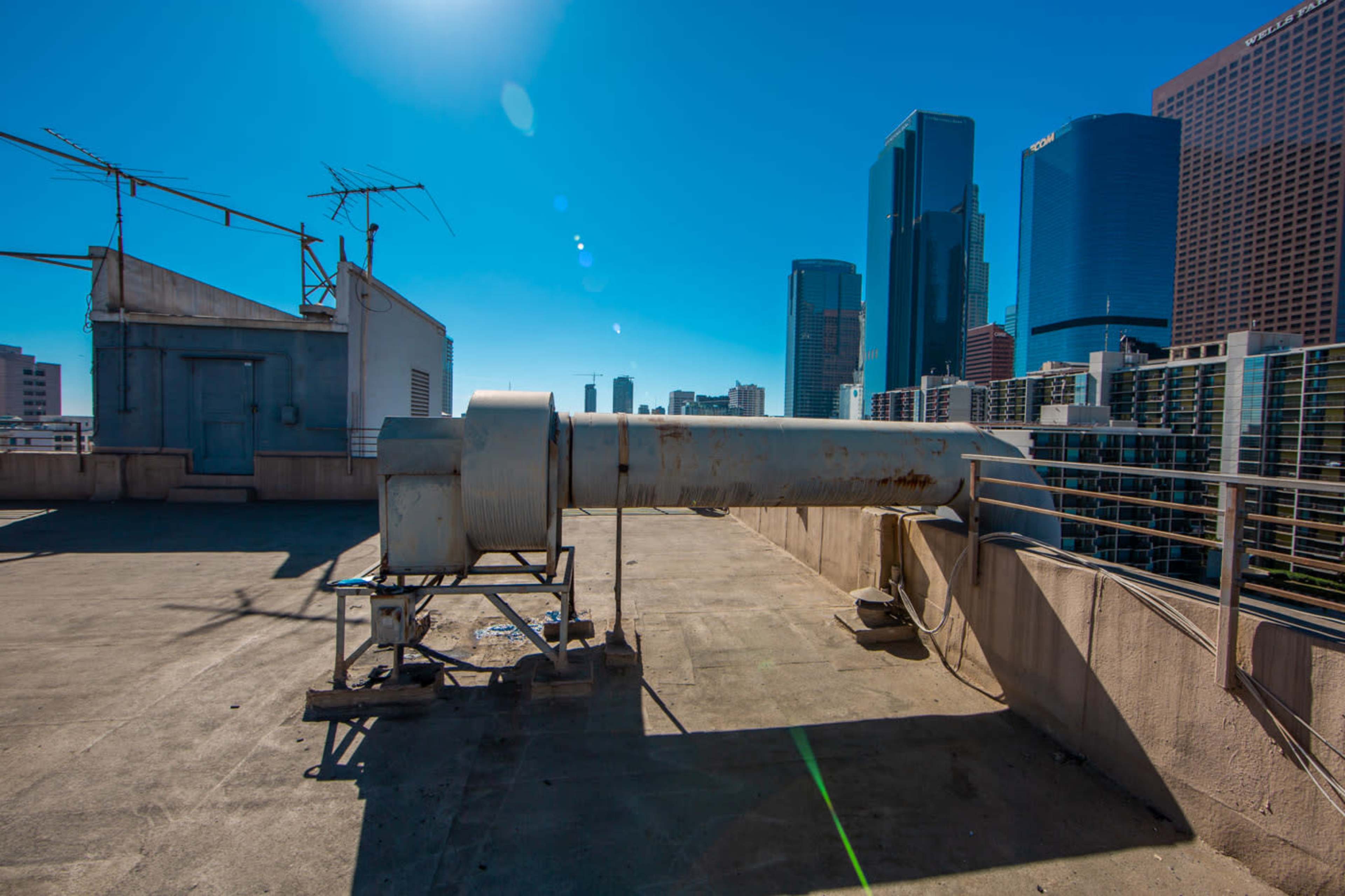 A large industrial vent pipe stands on a rooftop, with a skyline of modern skyscrapers visible in the background under a clear blue sky.