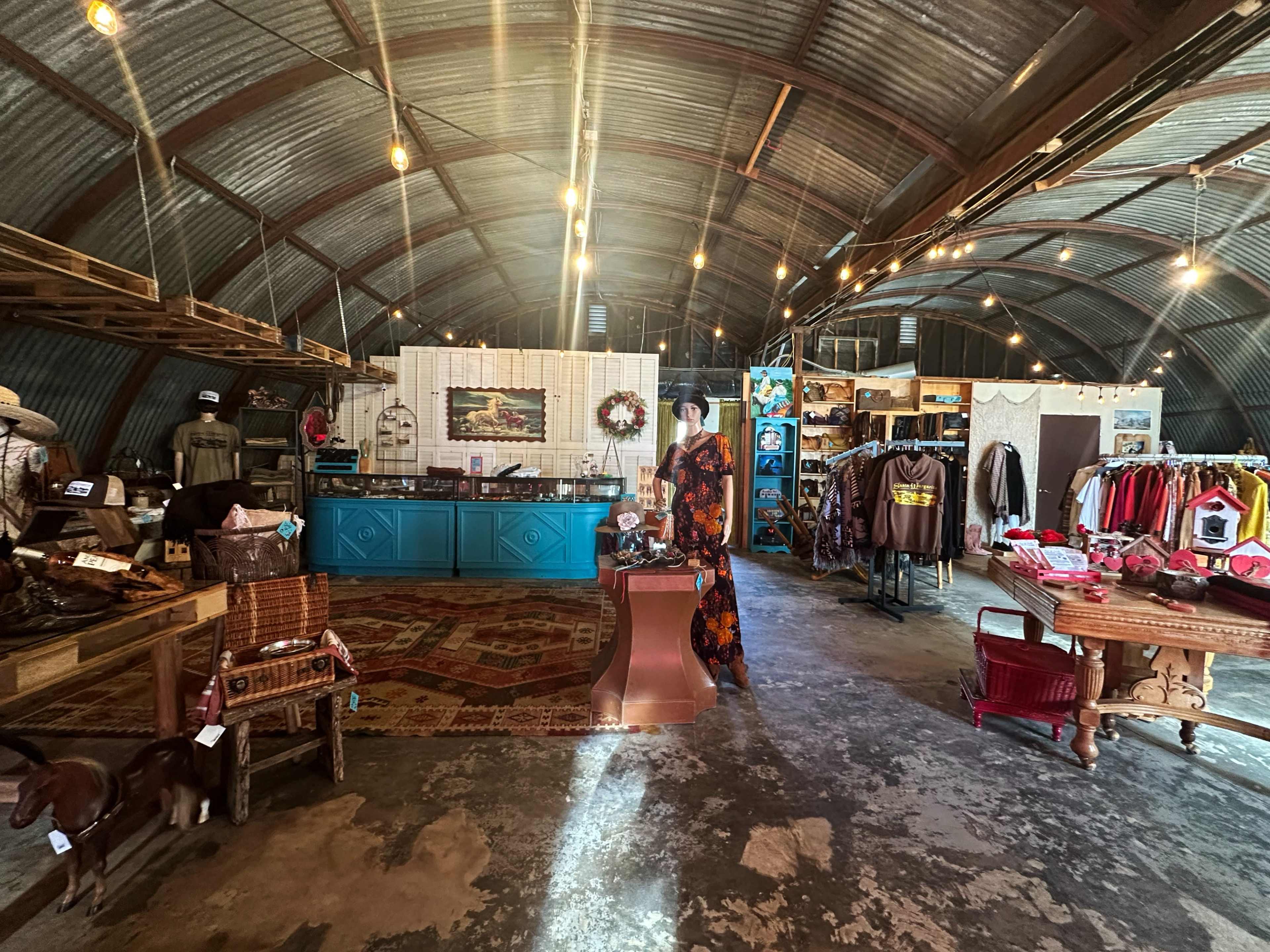 The interior of a rustic retail shop features wooden shelves, a turquoise counter, and various clothing and decor items arranged under string lights.