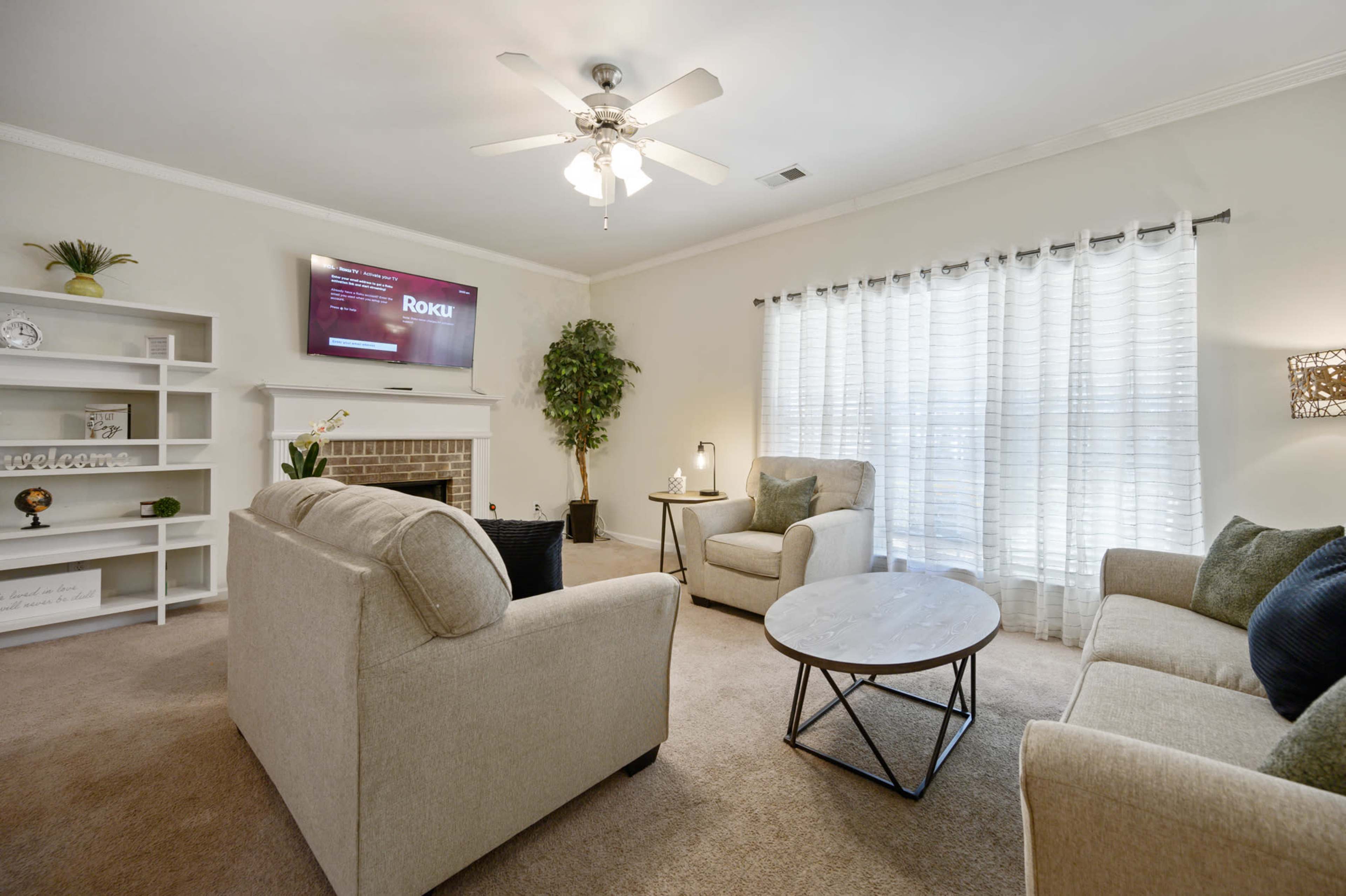 A cozy living room features two beige armchairs, a coffee table, a fireplace, and a wall-mounted TV, with light curtains allowing natural light to enter.