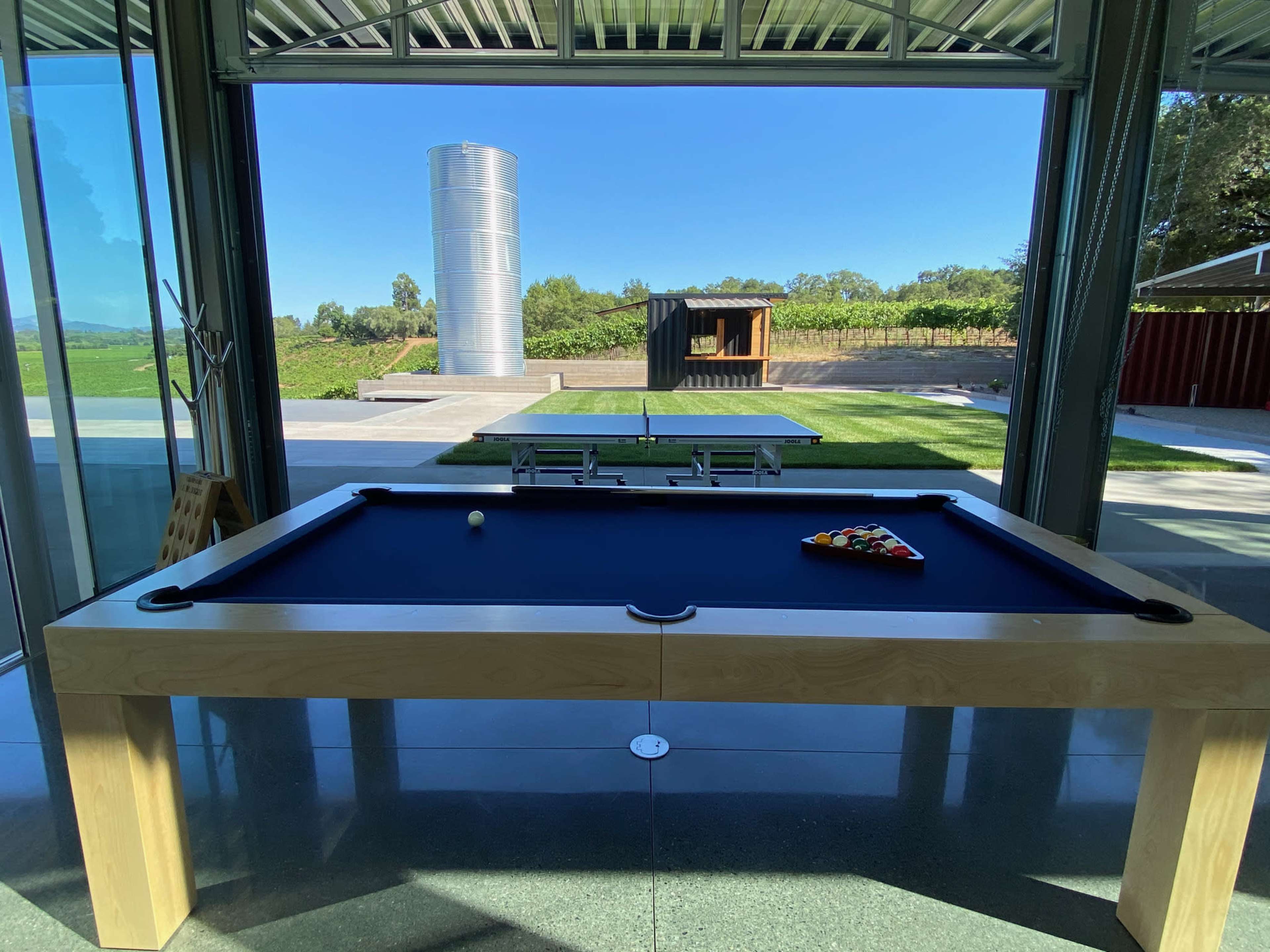 A pool table with a triangle of balls is positioned inside a modern building, facing a large glass window that overlooks a landscaped outdoor area.