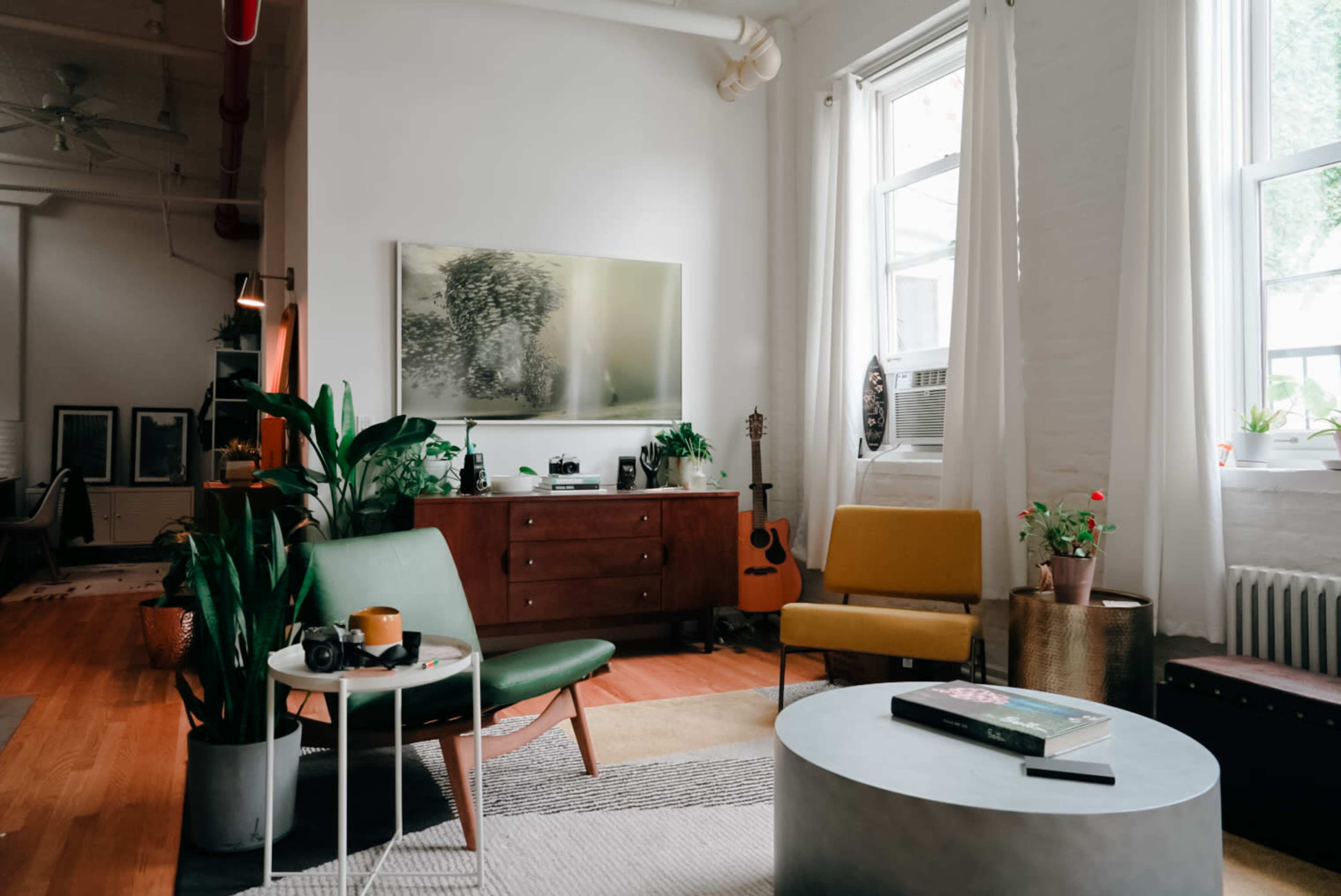 A bright living room features a green chair, a yellow chair, a coffee table, and a wooden cabinet, with a guitar leaning against the wall and plants in pots.