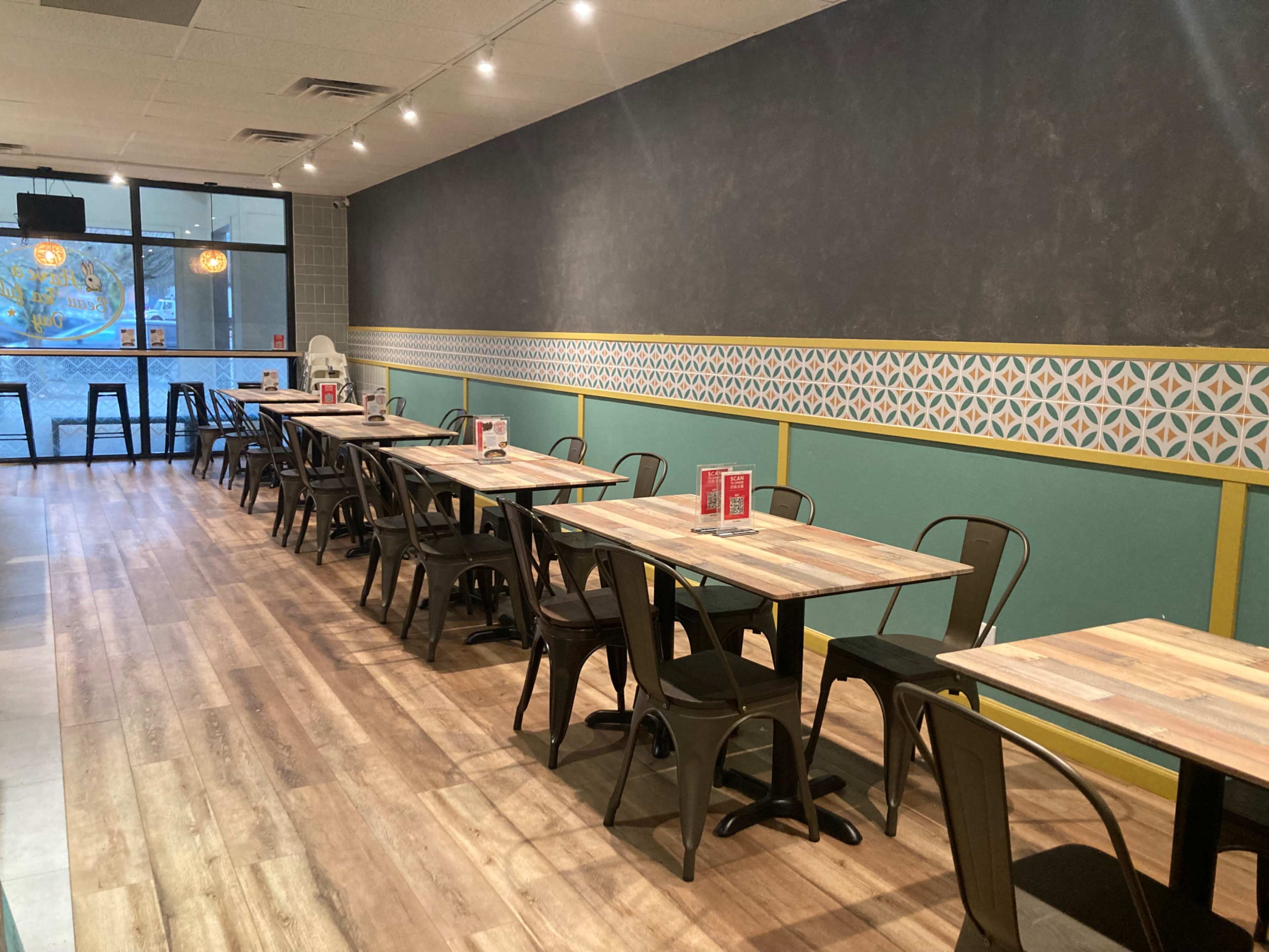 An empty restaurant with wooden tables and black chairs arranged in rows along a patterned wall.