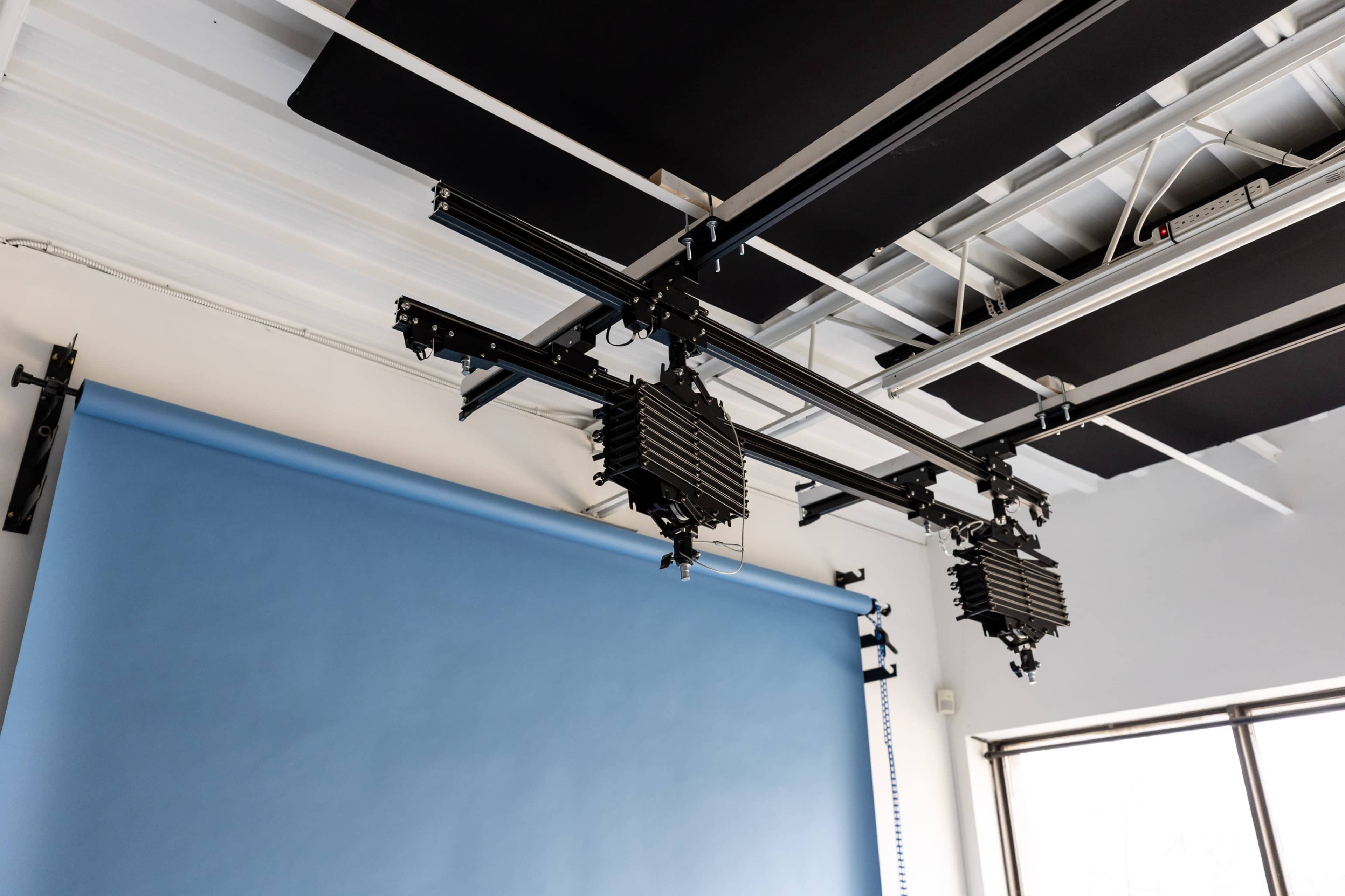 The image shows a photography studio setup featuring a blue backdrop and three suspended lighting fixtures mounted on a track system.