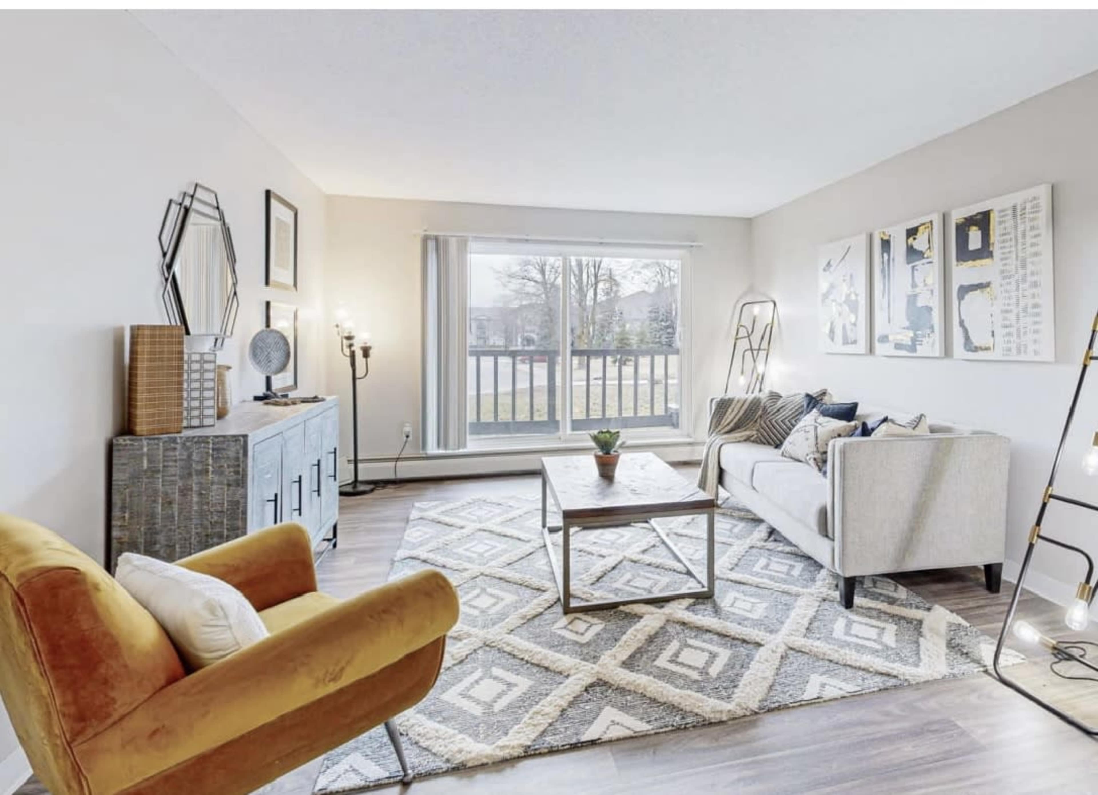 A modern living room features a light-colored sofa, an orange armchair, a coffee table, and a large window with a view of the outdoors.