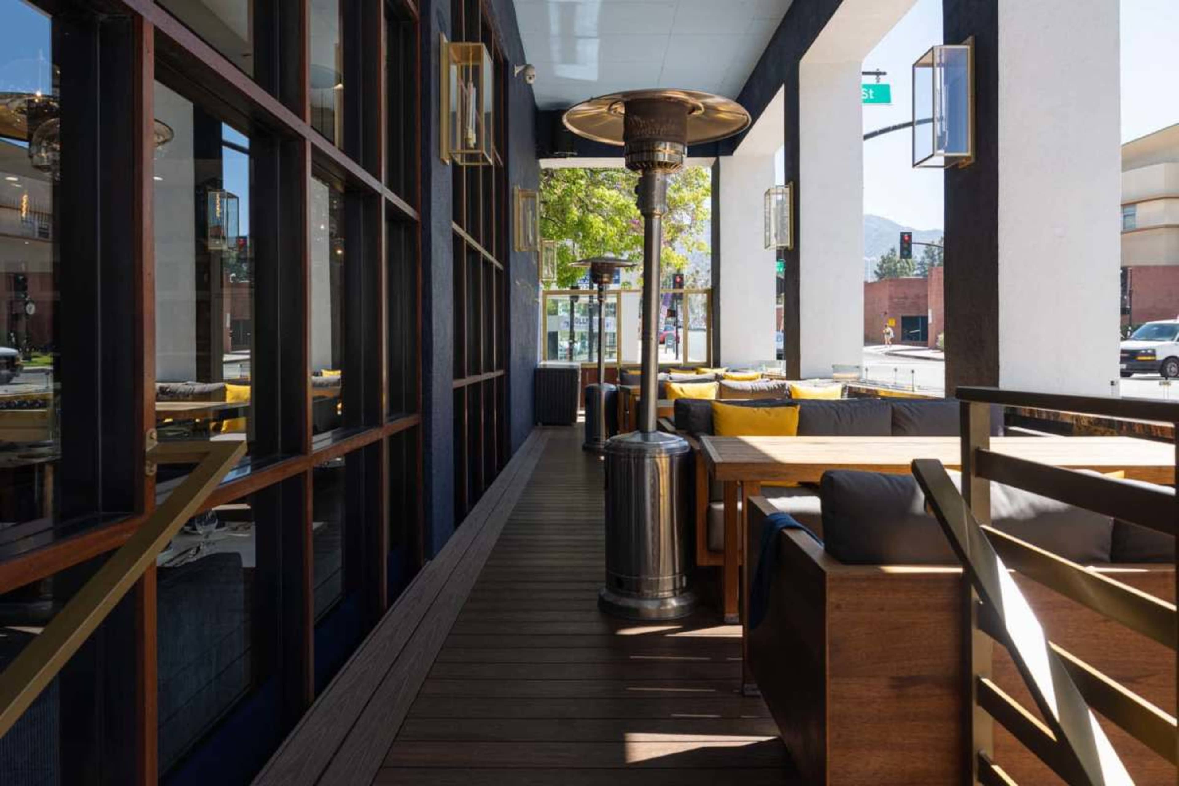 The image shows an outdoor dining area of a restaurant featuring wooden furniture, a patio heater, and large windows overlooking a street.