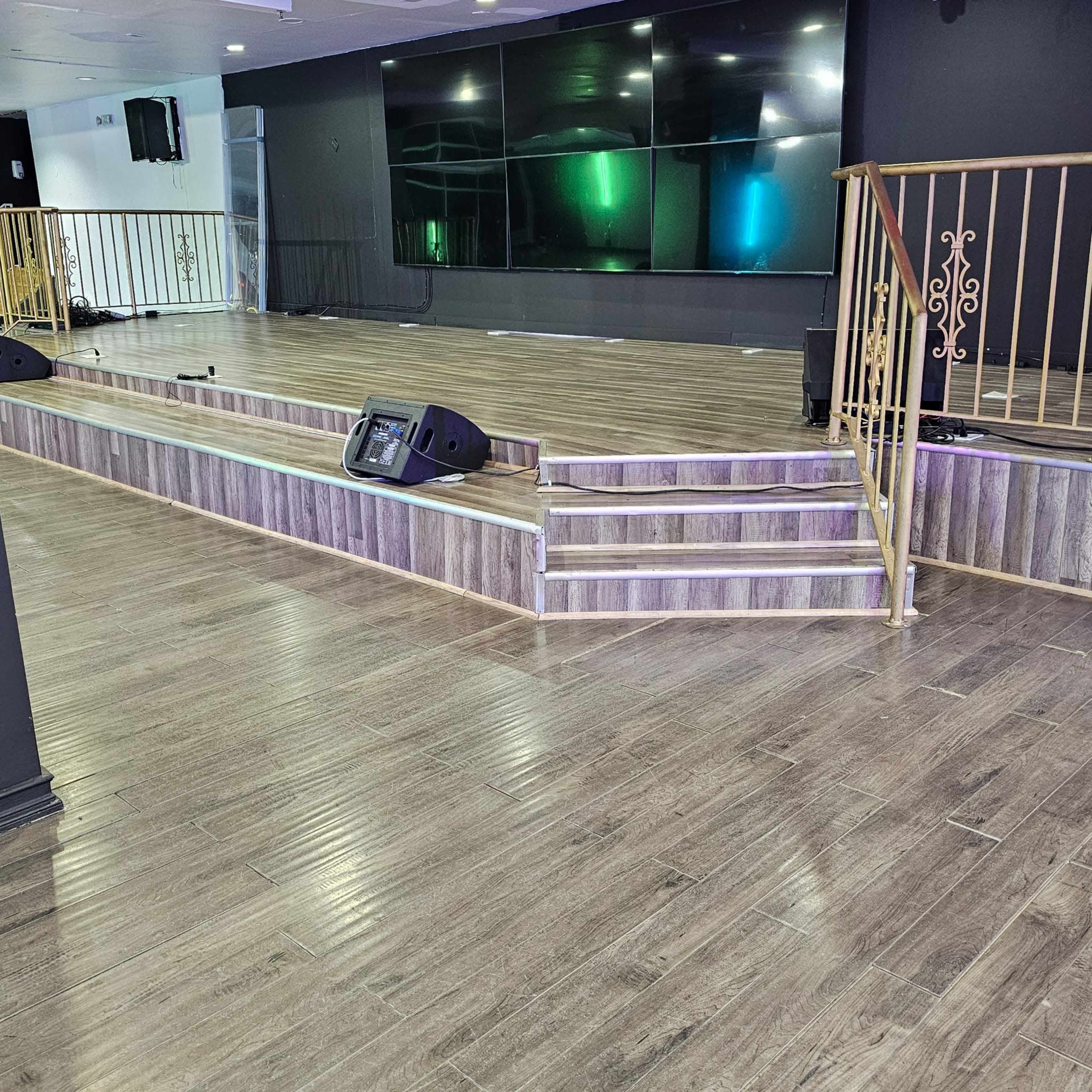 The image shows an indoor stage area with wooden flooring, stairs leading up to the stage, and large screens mounted on the wall.