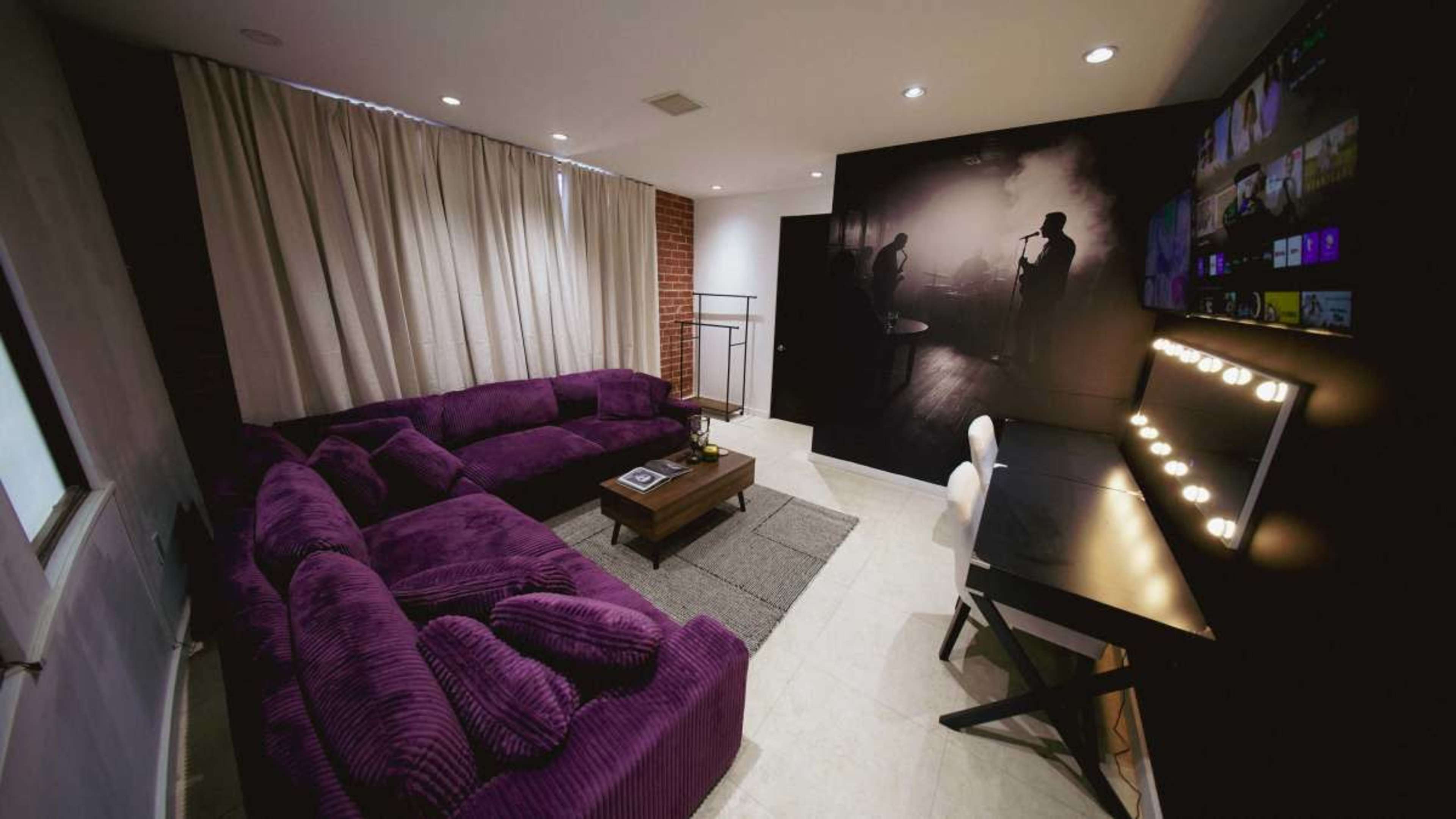 The image shows a living room with a purple sectional sofa, a wooden coffee table, and a desk with lights, surrounded by patterned curtains and a dark wall featuring a human silhouette.