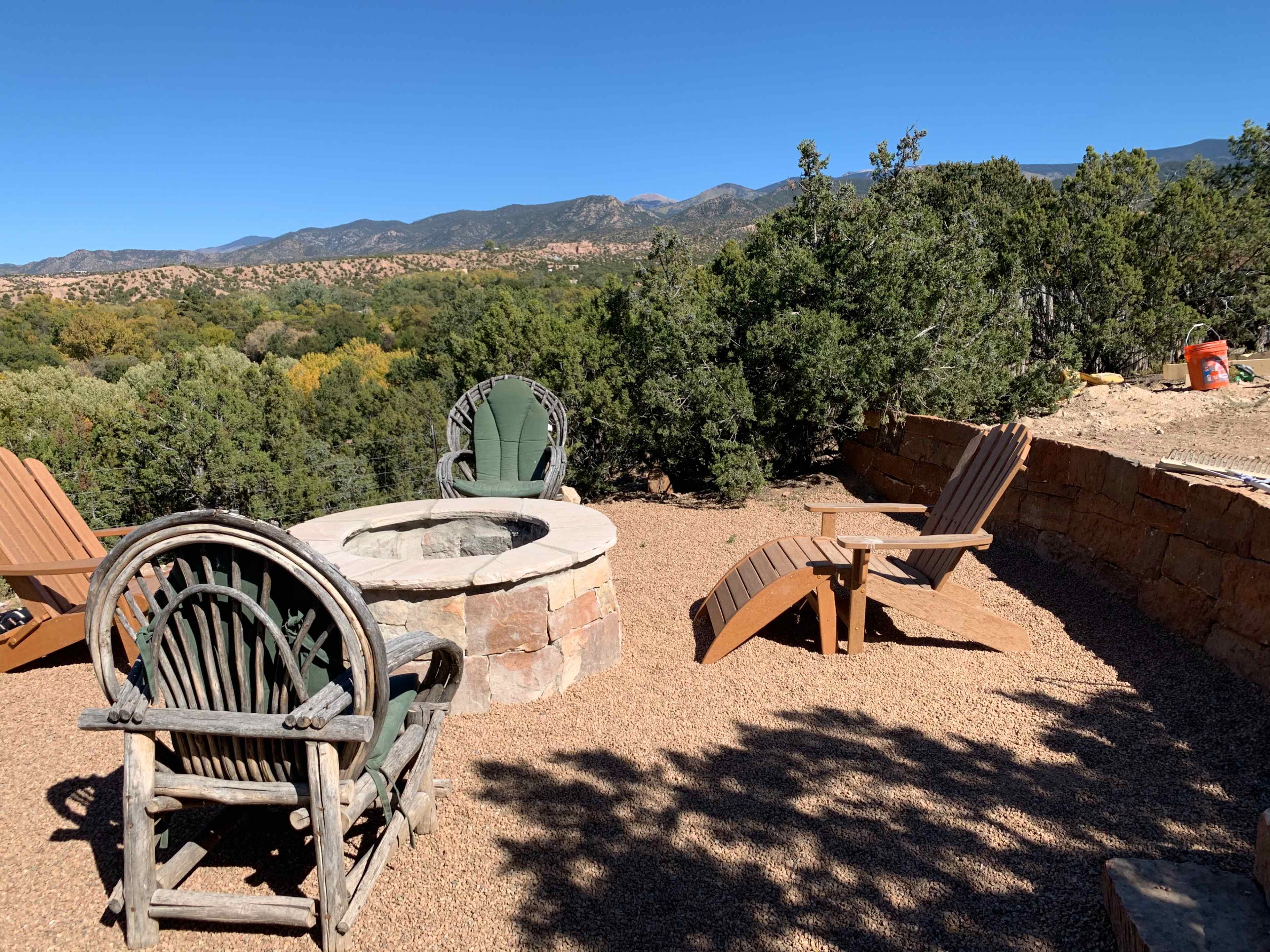 A stone fire pit is surrounded by wooden chairs and mountain greenery under a clear blue sky.