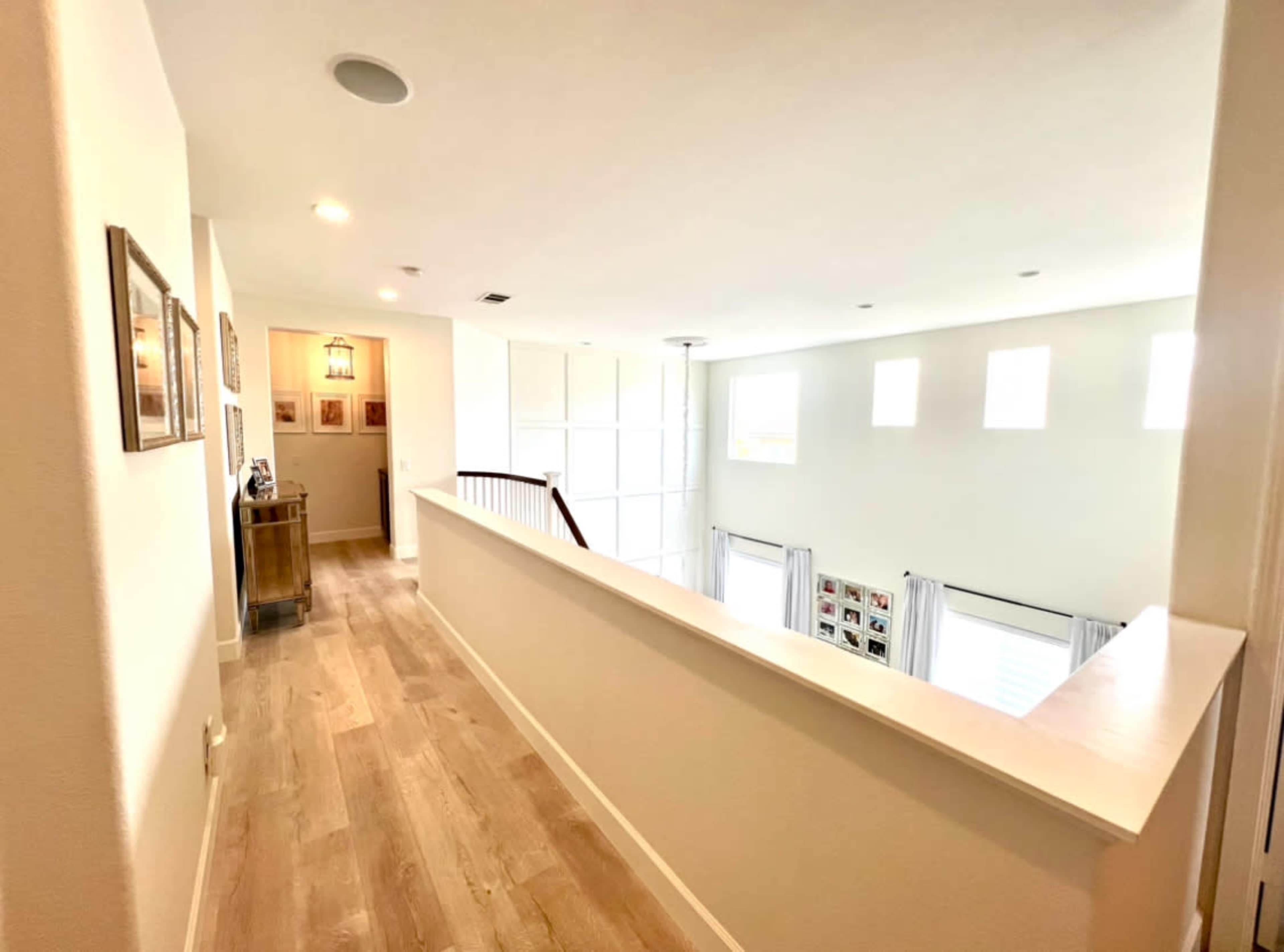 A bright hallway with a wooden floor leading to a balcony overlooking a spacious living area with large windows.