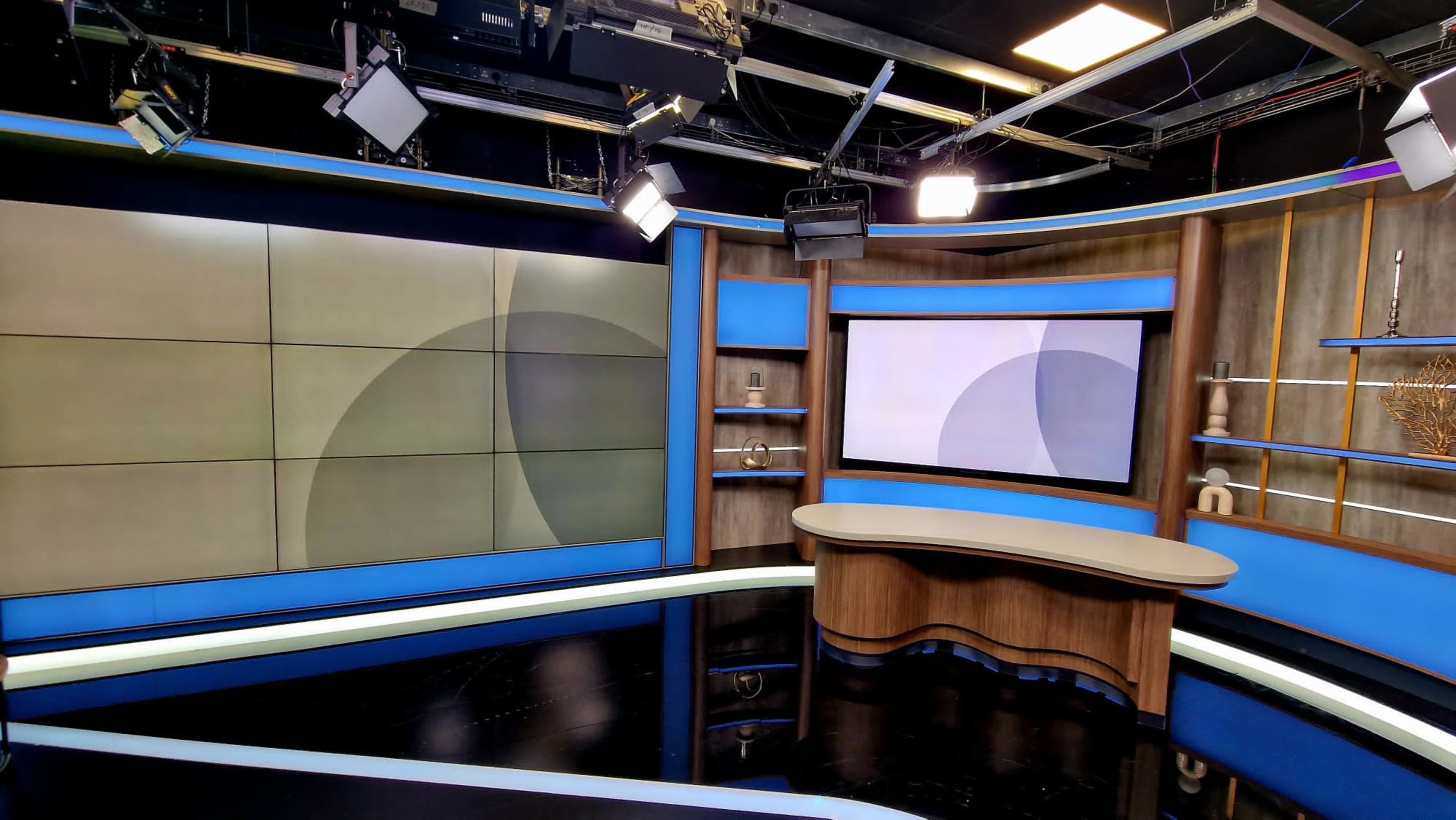 The image shows a modern television studio set with a large central desk, a curved screen, and multiple light fixtures overhead.