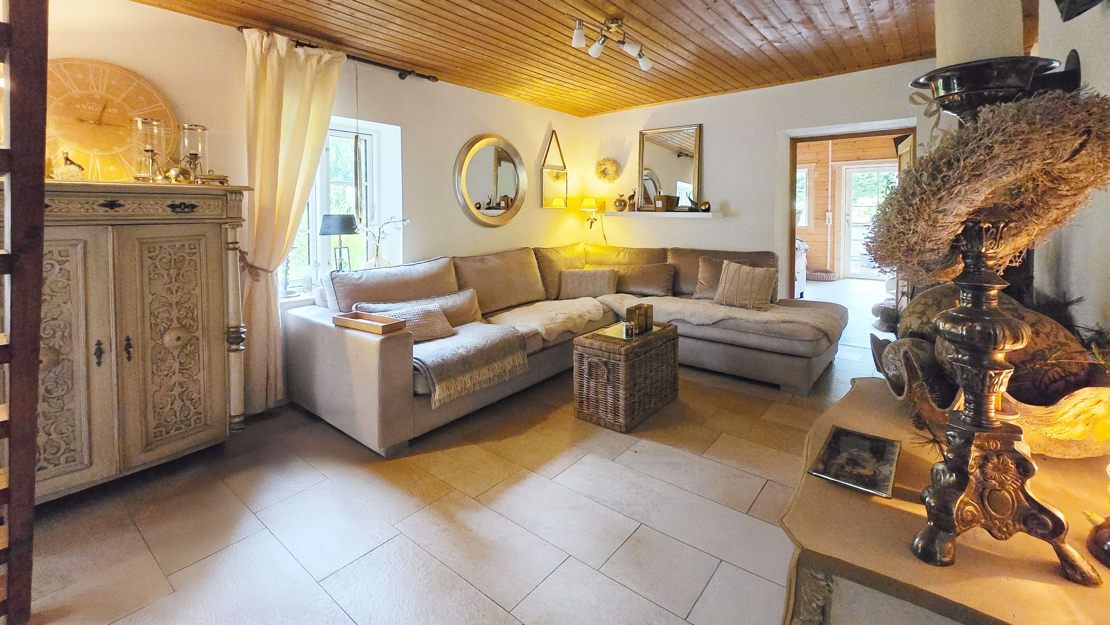 The image shows a cozy living room featuring a large sectional sofa, decorative mirrors on the walls, a wooden ceiling, and a wicker coffee table.