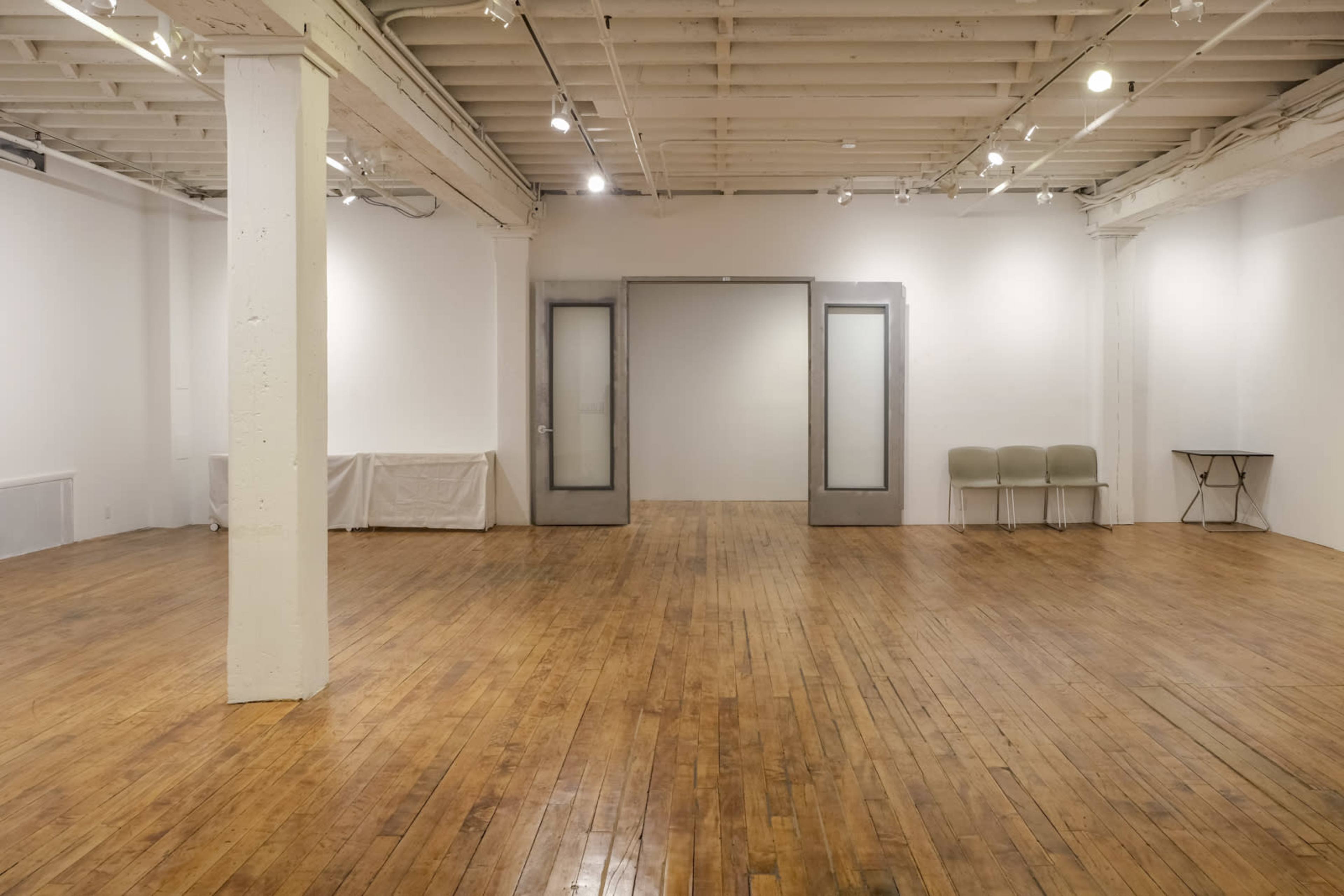 The image shows an empty, spacious room with wooden flooring, white walls, and a few pieces of furniture against the walls.