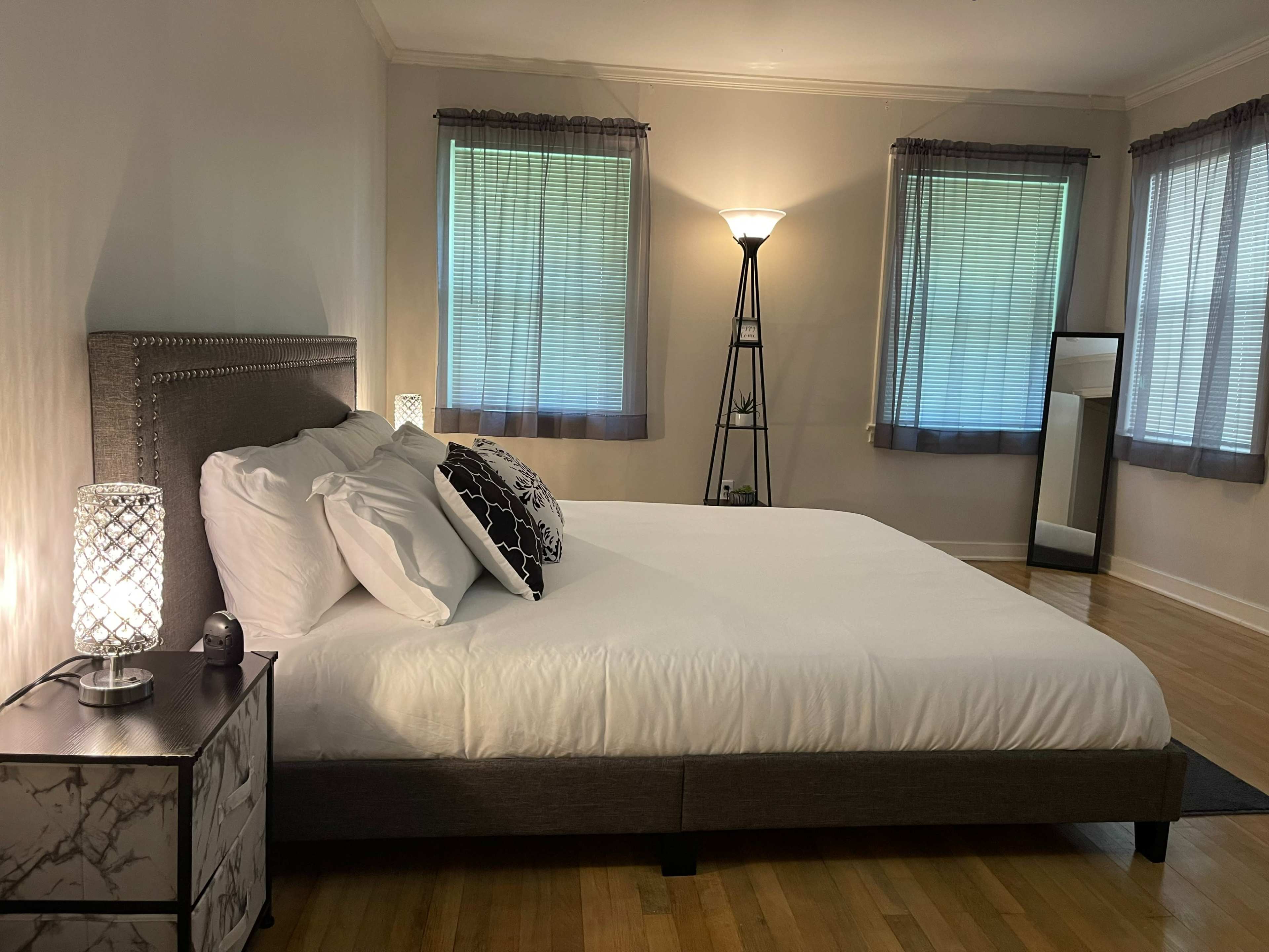 A spacious bedroom with a large bed, two bedside lamps, and tall windows dressed with sheer curtains.