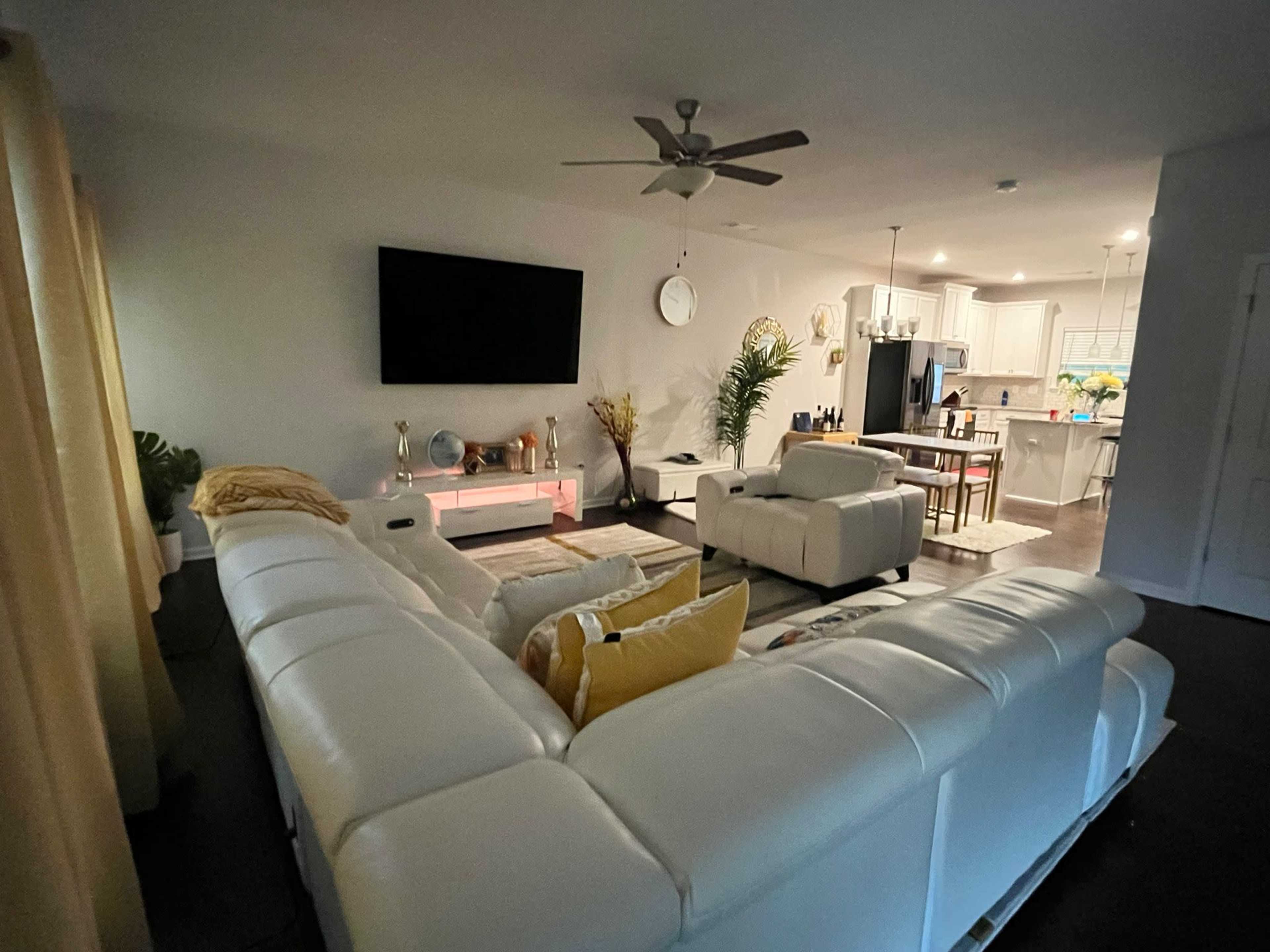 The image shows a modern living room with a white sectional sofa, a large wall-mounted television, and an open kitchen in the background.