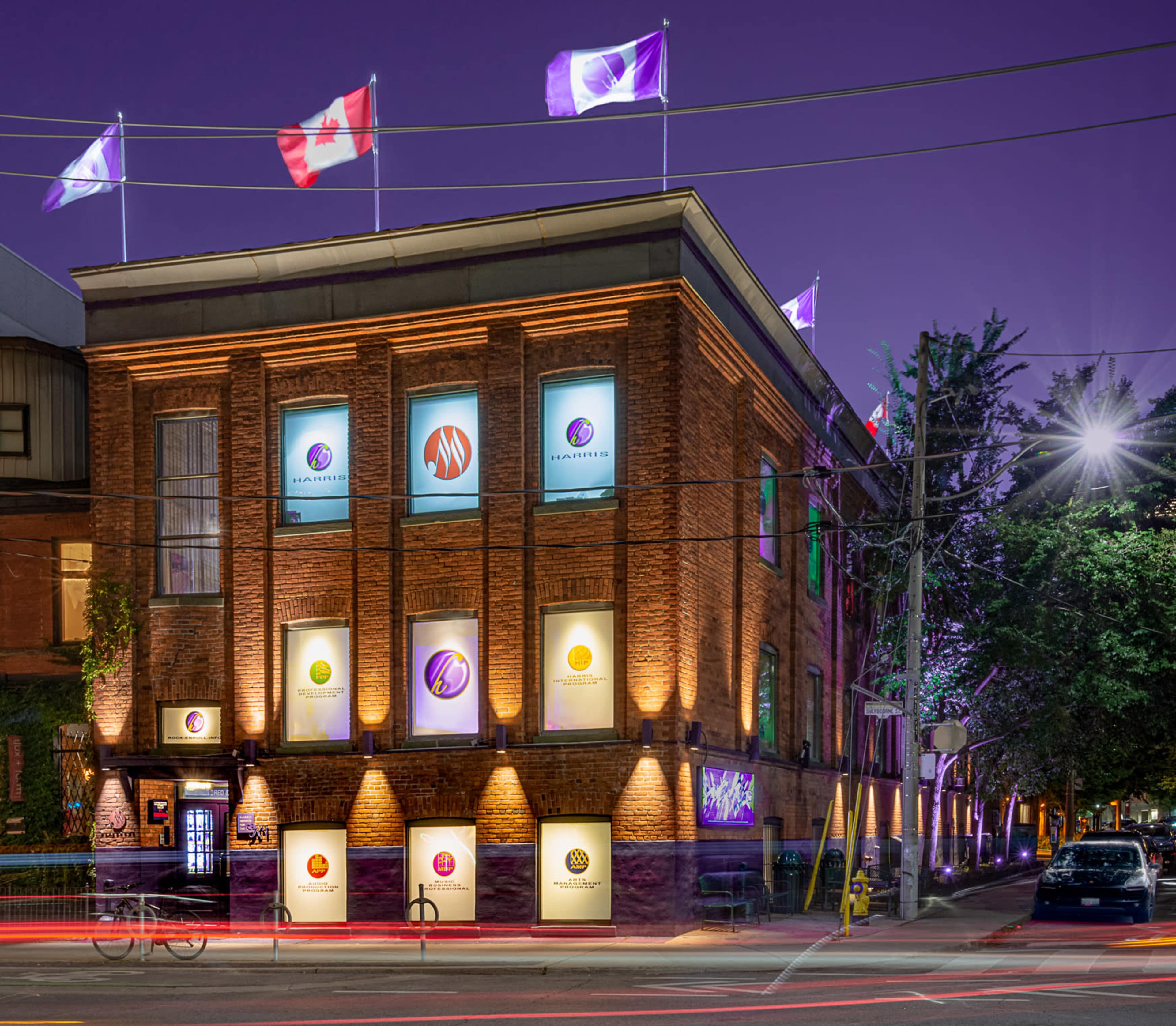 The image shows a brick building with illuminated windows, featuring design branding and multiple flags, including the Canadian flag, flying from its roof at dusk.