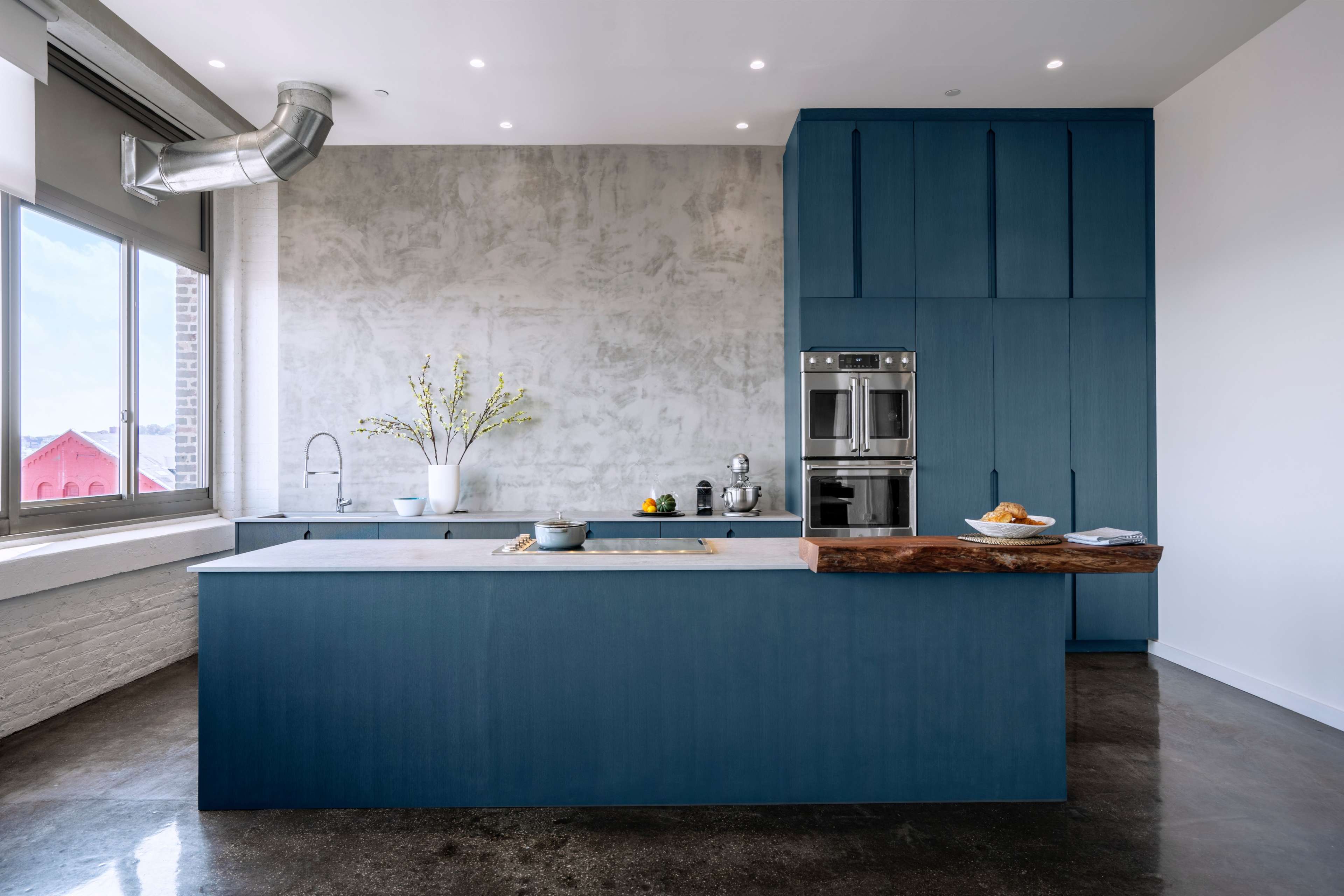 A modern kitchen featuring blue cabinetry, a large island with a wood countertop, and a concrete wall behind a window.