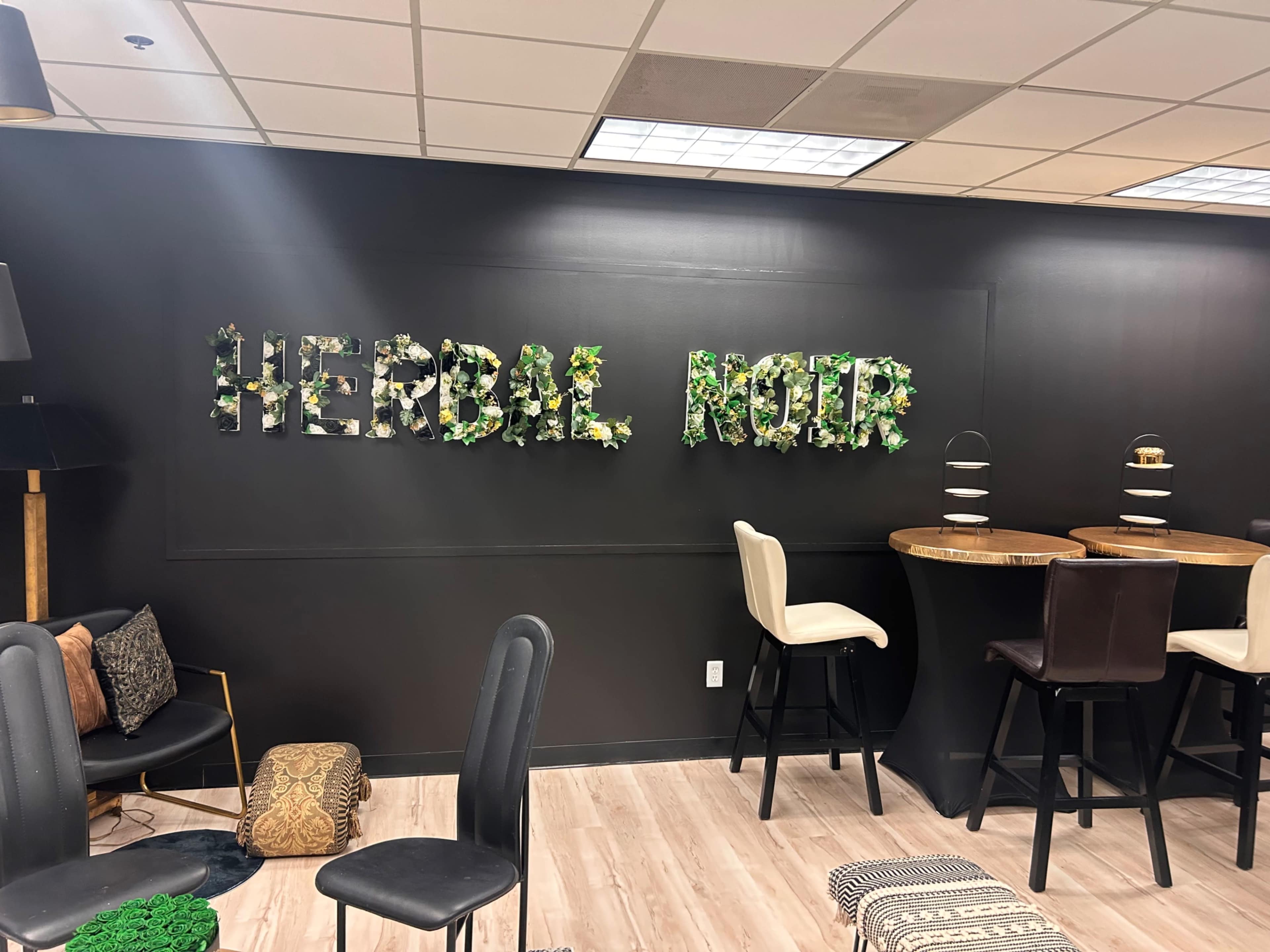The image shows a modern interior space featuring a black wall with the words "HERBAL NOIR" decorated with greenery, alongside a dining area with wooden tables and black chairs.