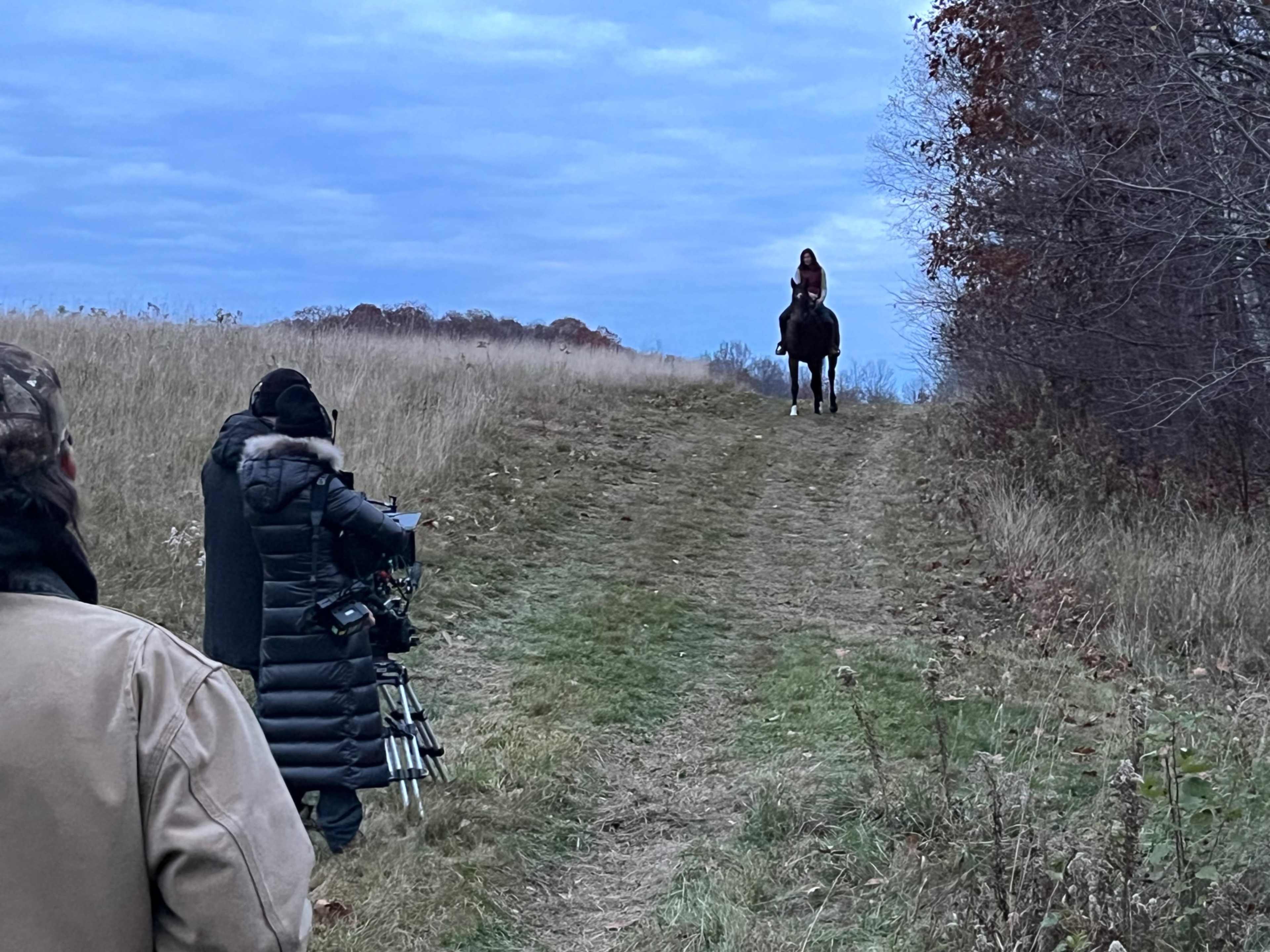 A woman rides a horse along a grassy path while a film crew sets up equipment in the foreground.