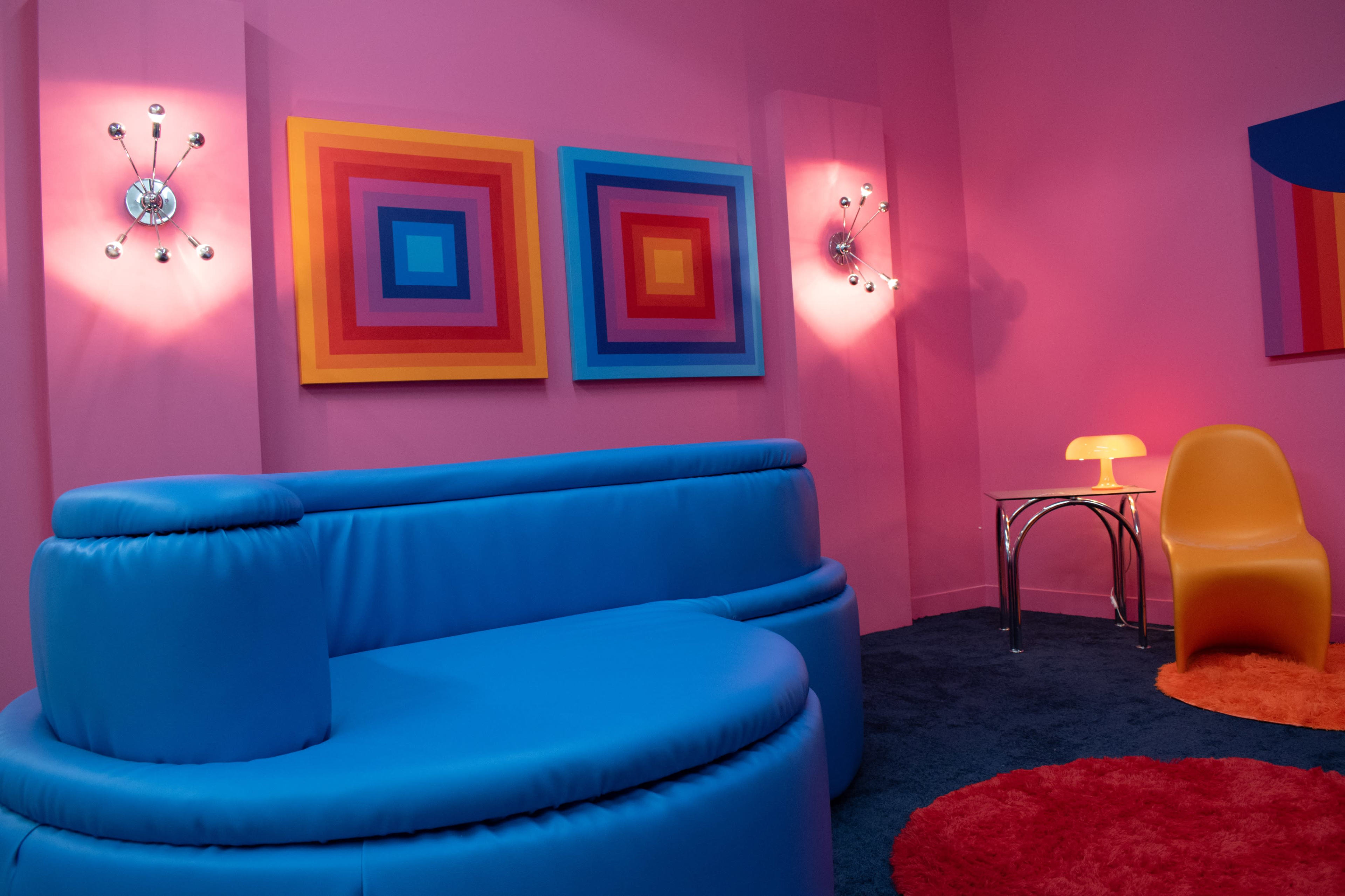 A brightly colored room features a blue circular couch, an orange circular rug, and two abstract art pieces on the pink walls.