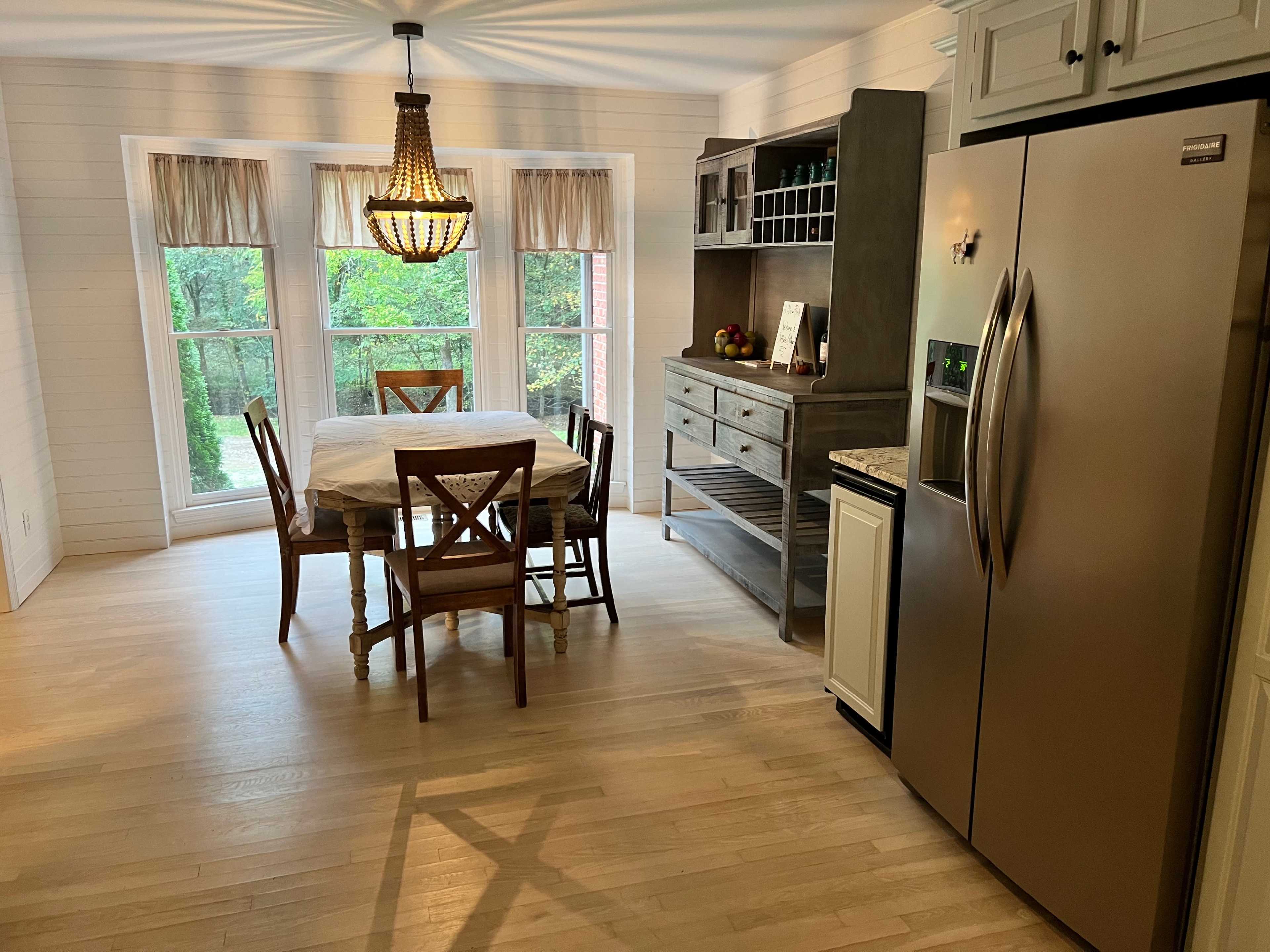 A dining area features a wooden table with four chairs, a sideboard, and large windows overlooking a green landscape.