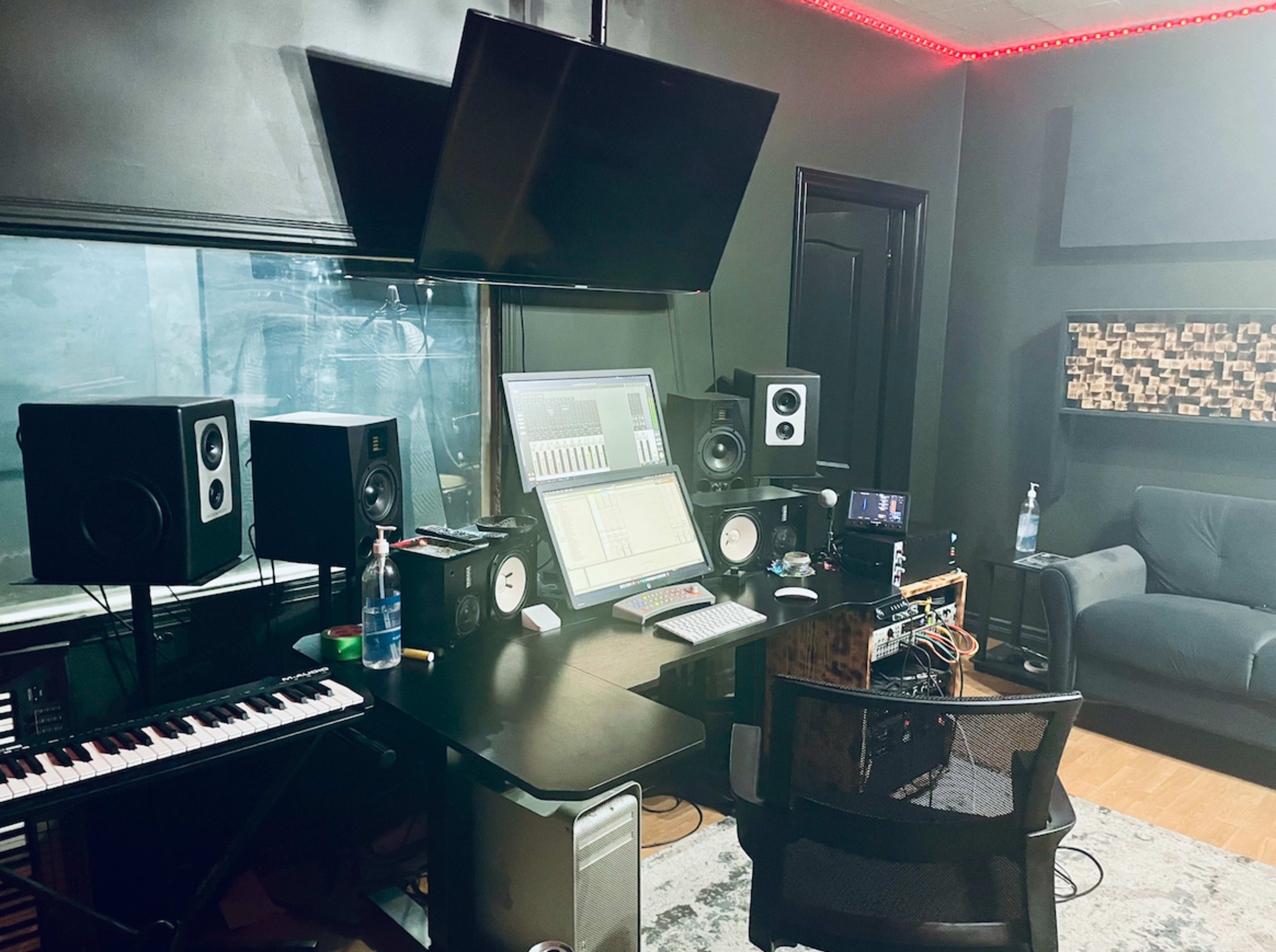 The image depicts a music production studio featuring a computer workstation, speakers, a keyboard, and a cozy seating area.