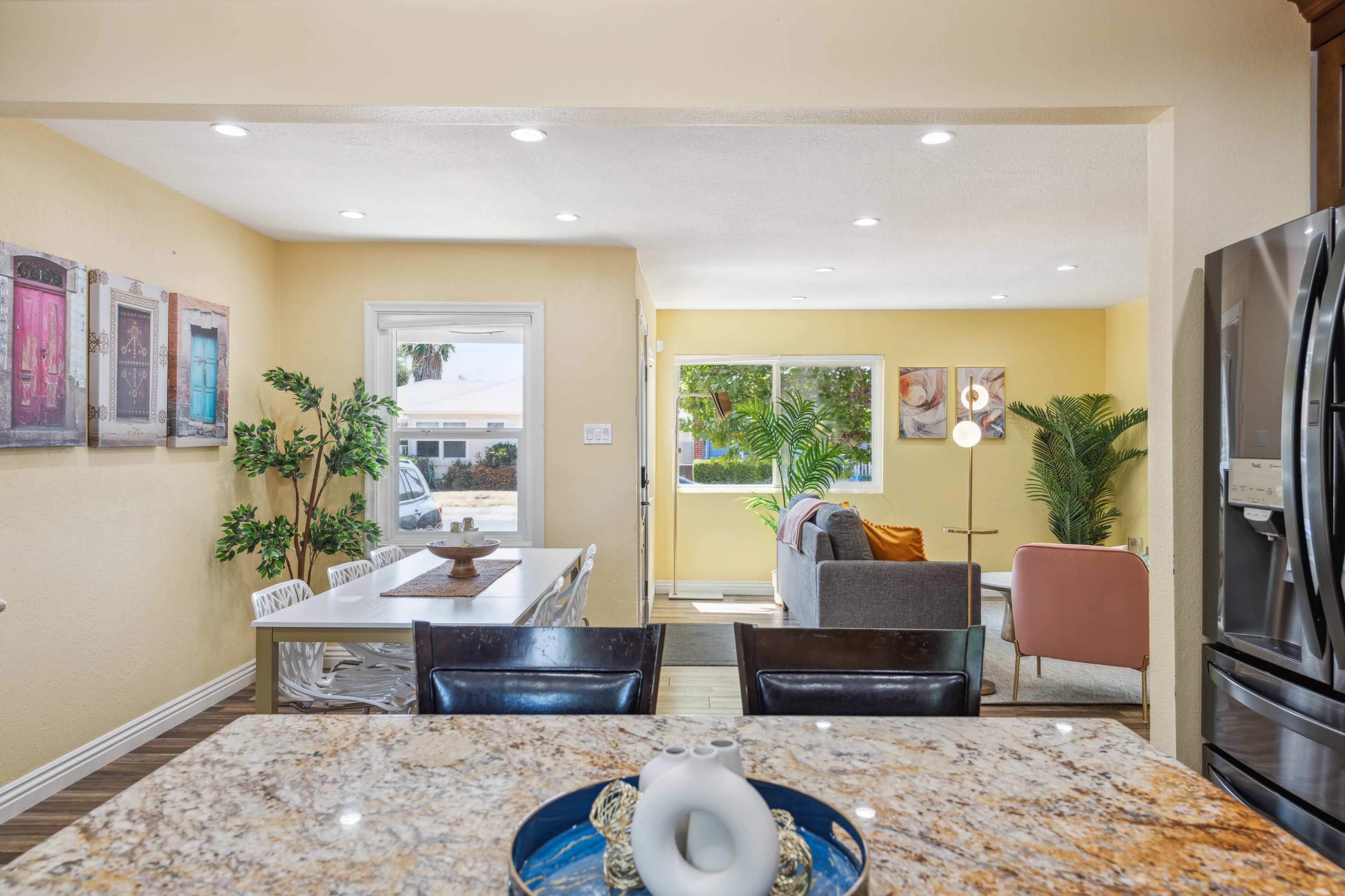 A brightly lit open-concept living space featuring a dining area with a long table, a kitchen with granite countertops, and a cozy living room furnished with a gray sofa and an armchair.
